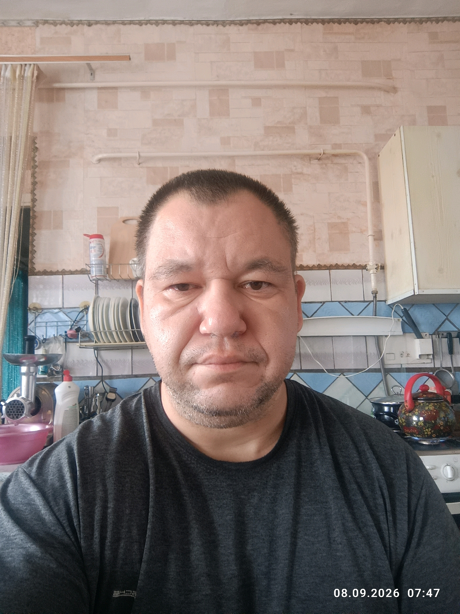 A head-and-shoulders, frontal, selfie-style photograph shows a middle-aged man with a light complexion and short dark hair. He is wearing a dark grey t-shirt and is looking directly at the camera with a neutral expression. The background appears to be a kitchen. Visible objects include kitchen cabinets, shelves with dishes and bottles, a meat grinder, and a decorative teapot. The timestamp "08.09.2026 07:47" is superimposed in the lower right corner of the image. The location specified is Kanovo, Russia.