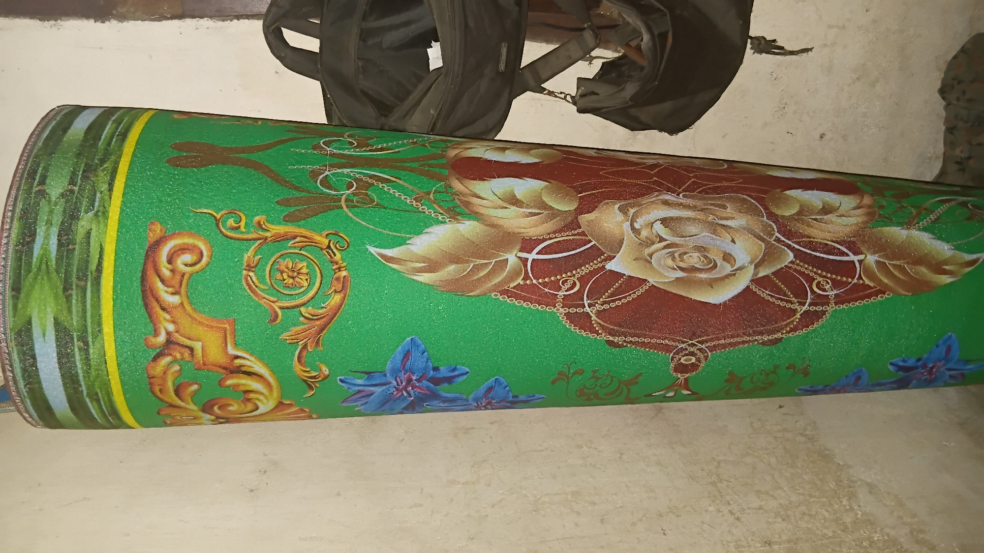A green roll of material, possibly a rug or mat, is leaning against a textured off-white wall. The material has a textured surface and is decorated with a repeating pattern of ornate gold scrollwork, blue flowers, and a large stylized rose in shades of red and gold. A yellow stripe runs along the top edge of the roll. To the right of the roll, two black bags are hanging from an unseen support, one larger bag above a smaller one. The background wall is rough and shows signs of age. The scene appears to be indoors, possibly in a storage area or a less formal part of a building.