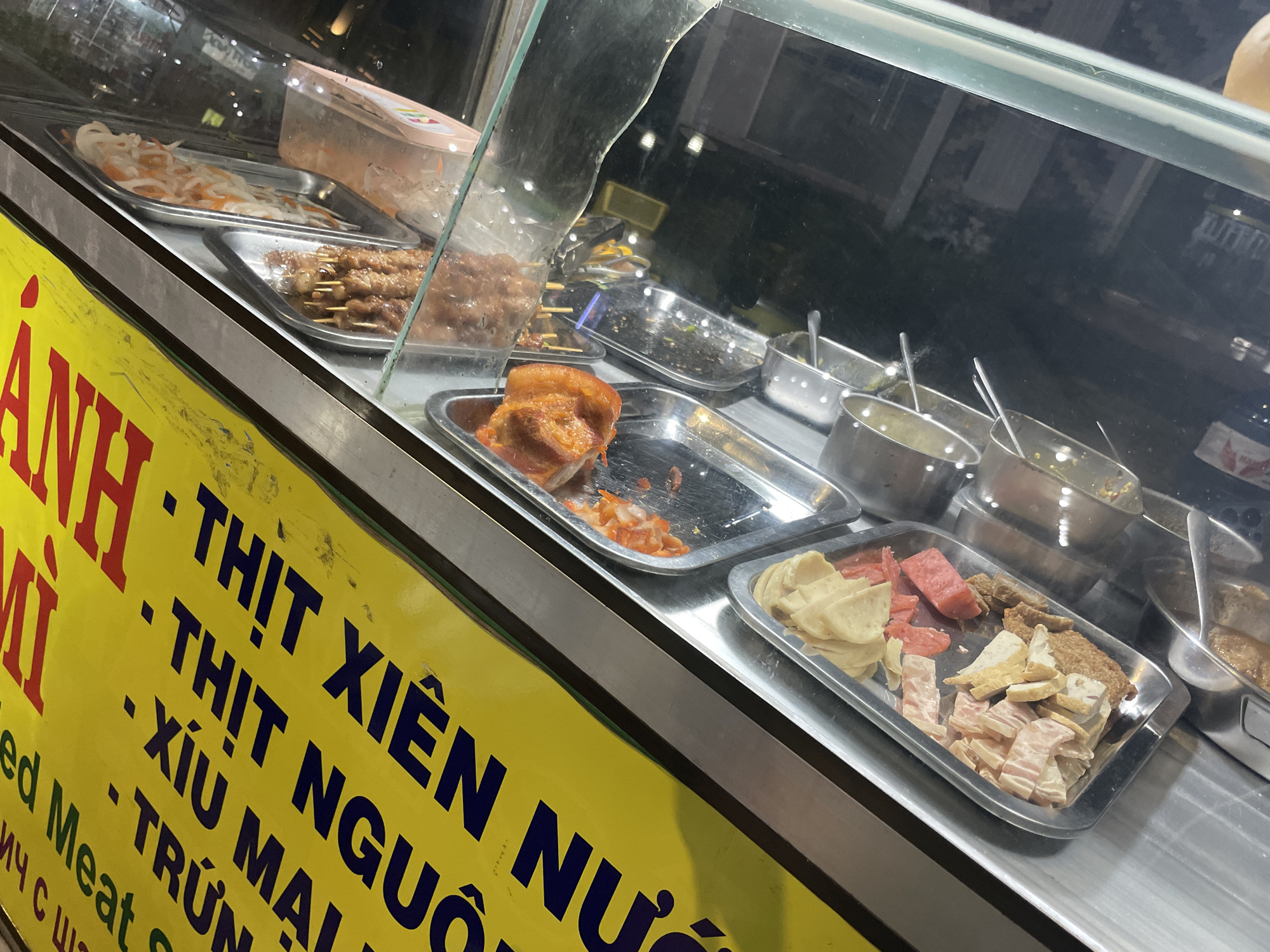 This image depicts a Vietnamese street food stall, situated in Mũi Né, Vietnam, operating at night. The stall features a glass-fronted display case housing various prepared ingredients. A prominent yellow sign on the left advertises "BÁNH MÌ" (Banh Mi) and lists available fillings, including "THỊT XIÊN" (grilled pork skewers), "THỊT NGUỘI" (cold cuts), "XÍU MẠI" (meatballs), and "TRỨNG" (egg). Inside the display, multiple stainless steel trays contain food items: one tray holds white and orange pickled vegetables (likely daikon and carrots); another contains several grilled pork skewers; a third displays a large piece of roasted pork belly; and a fourth tray presents an assortment of sliced cold cuts and processed meats, some with a reddish hue. Several smaller metal bowls filled with various sauces or condiments are also visible behind the food trays. The scene is illuminated by artificial light, reflecting off the glass and metal surfaces, indicating an outdoor or semi-outdoor commercial setting. No individuals are present in the frame.