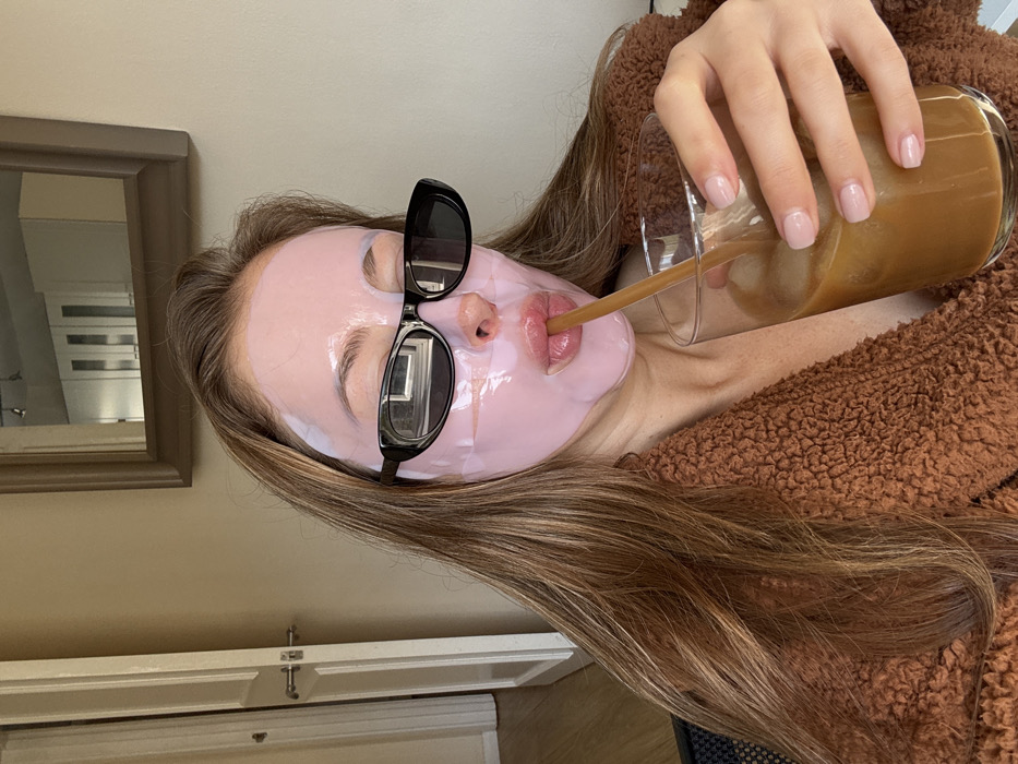 An individual, identified as female, is depicted indoors from the chest up, wearing a translucent pink sheet face mask that covers her facial skin, excluding her lips and the areas behind her black, oval-framed sunglasses. Her long, light brown hair is visible around her face. She is wearing a brown, textured, sherpa-style garment. Her right hand, featuring light pink manicured fingernails, holds a clear glass containing an iced, light brown beverage, into which a brown straw is inserted and positioned at her lips, indicating she is drinking. The background features a light-colored wall. To the left, a white door with a silver handle is partially visible. Centered behind the individual, a framed mirror reflects an overhead light fixture and light-colored cabinetry. The specific location in the city of Unknown, Unknown is an indeterminate indoor setting.