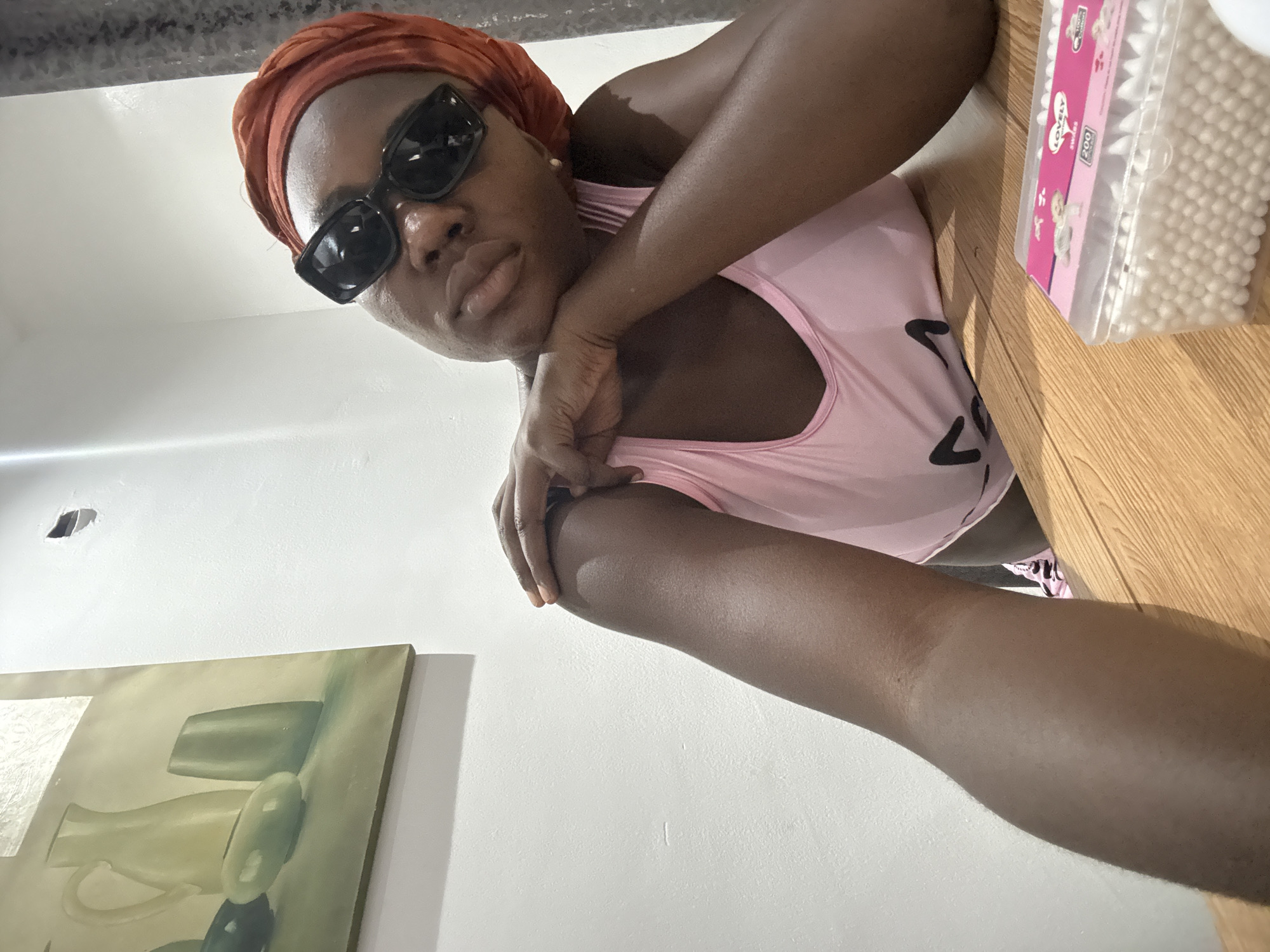 This media file presents a rotated, low-angle indoor shot primarily featuring a woman. Her head is positioned on the left side of the frame, with her body extending diagonally towards the right.
The central subject is a woman with dark skin, wearing a rust-colored headwrap and black rectangular sunglasses that obscure her eyes. She is dressed in a light pink tank top. Her lips are slightly parted in a subtle pout, and her left hand is positioned near her chin or neck. Her arms are prominently featured in the foreground, close to the camera.
The background consists mainly of a plain white wall. In the upper right portion of the image, a wooden surface, likely a shelf or table, holds a clear plastic container filled with cotton swabs. A framed painting or piece of artwork, depicting a still life with muted green and gray forms resembling vases or bottles, is visible on the lower left section of the wall.
The plastic container of cotton swabs features a pink label with the text "LOVELY SWABS" and "200" prominently displayed, along with small cartoon baby illustrations. The lighting appears to be natural indoor light. The woman's expression conveys a sense of confidence or seriousness as she looks directly at the viewer.