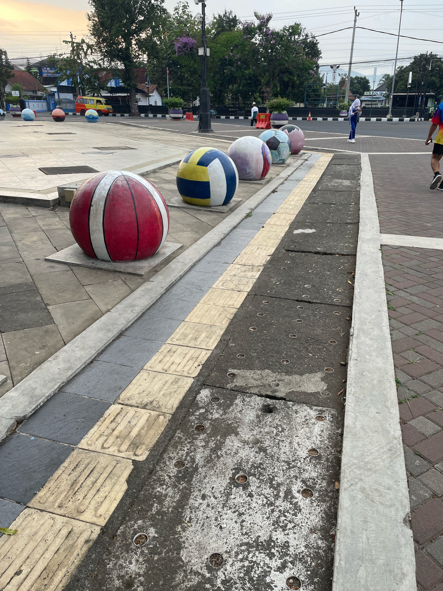 The image captures an outdoor scene on a cloudy day, likely in the city of Purworejo, Indonesia, given the context. In the foreground, a row of large, decorative spherical objects resembling exercise balls are arranged along a paved pathway. The closest ball is red and white striped, followed by a blue, yellow, and white patterned ball, and then several more balls with varying color schemes, including some with blue, white, and pink accents. The pathway features tactile paving, indicated by the textured yellow tiles next to gray square tiles, suggesting a pedestrian area.
Beyond the pathway, a street is visible with a yellow and orange van in the background. Further back, buildings and trees line the street, and a light pole with a camera and what appears to be a spotlight is prominent. A few individuals are present in the scene. To the right, a person in athletic wear, including a colorful t-shirt and shorts, is walking away from the camera. In the middle ground, a person in a white hijab and blue athletic attire is standing and looking down at something in their hands. Another person in a white shirt and dark pants is walking in the distance. The overall environment suggests a public space, possibly a park or a plaza, adjacent to a street in an urban setting. The ground surfaces include concrete, paving stones, and asphalt.