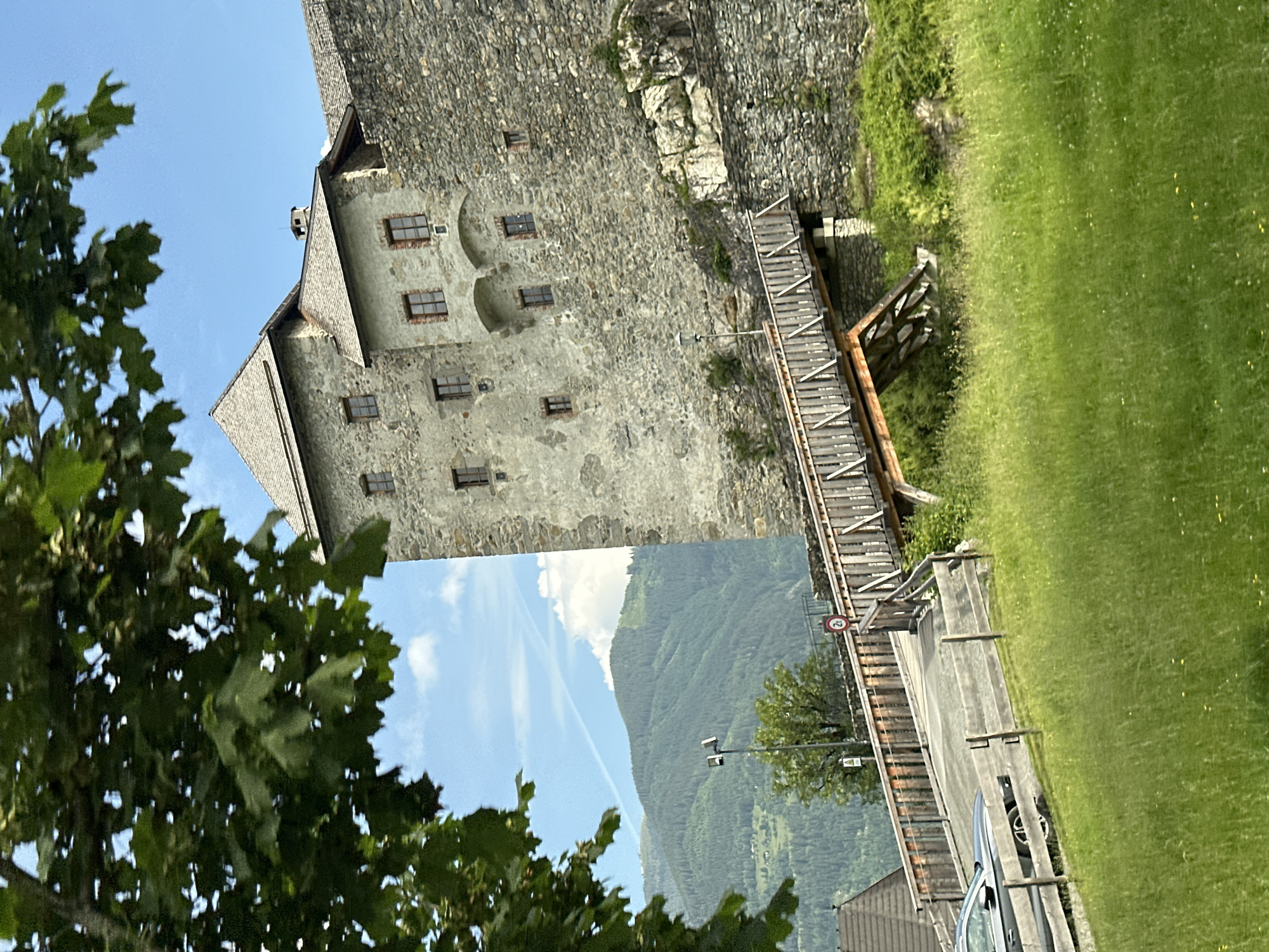 The image shows a section of Kaprun Castle, also known as Burg Kaprun, located in Kaprun, Austria. The castle is constructed of stone and features multiple towers with grey tiled roofs. A wooden bridge with railings spans across a section of the castle grounds, leading towards the stone structure. In the foreground, a grassy hill slopes upwards, with a fence and a section of a road visible. A car is parked on the road behind the fence. In the background, lush green mountains dominate the landscape under a bright blue sky with scattered clouds. A single streetlight stands on the grassy area near the bridge.