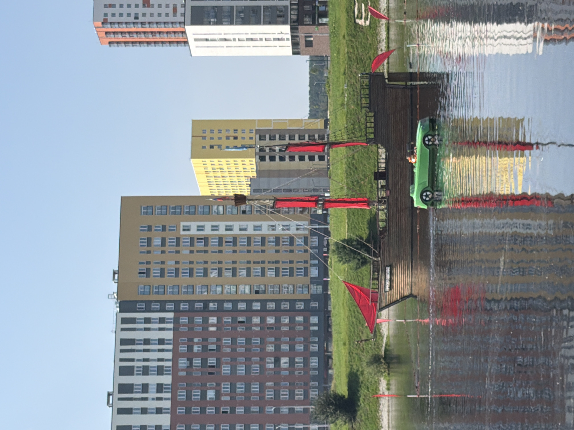 The image depicts a scenic waterfront with modern apartment buildings in the background and a pirate ship replica on the water in the foreground. The buildings are tall and colorful, with a mix of red, yellow, white, and brown facades, suggesting a contemporary urban development. One of the buildings has a distinctive orange and white exterior. In the middle ground, a grassy embankment separates the water from the buildings.
The focal point of the foreground is a wooden pirate ship replica, complete with masts and red sails. The ship appears to be a decorative element or a recreational attraction. Alongside the ship, a small, bright green toy boat with two figures inside is visible on the water. These figures appear to be children wearing life jackets, indicating a recreational activity. The water reflects the colors of the buildings and the ship, creating a shimmering effect.
The environment is bathed in sunlight under a clear blue sky, suggesting a pleasant day. The presence of multiple residential buildings indicates an urban or suburban setting. The specific location is the embankment of the Iset River in Yekaterinburg, Russia, specifically near the residential complex "Sky City." The architectural style of the buildings and the overall landscape are characteristic of modern Russian urban development projects.