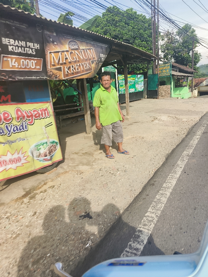 A man in a light green shirt and khaki shorts stands on a dirt road in front of a food stand. He is looking at the camera and has his hands on his hips. The food stand has a large sign that says "Magnum Kretek" and a smaller sign that says "Berani Pilih Kualitas" with a price of "14.000.-". There is a picture of a bowl of food on the stand as well. The man is standing on the edge of a paved road, which has a white stripe down the middle. There are trees and a small building on the other side of the road. The location is Jatiluhur, Indonesia, based on the coordinates (latitude: -6.571, longitude: 107.44). The photo is taken from a vehicle looking at the food stand and the man.