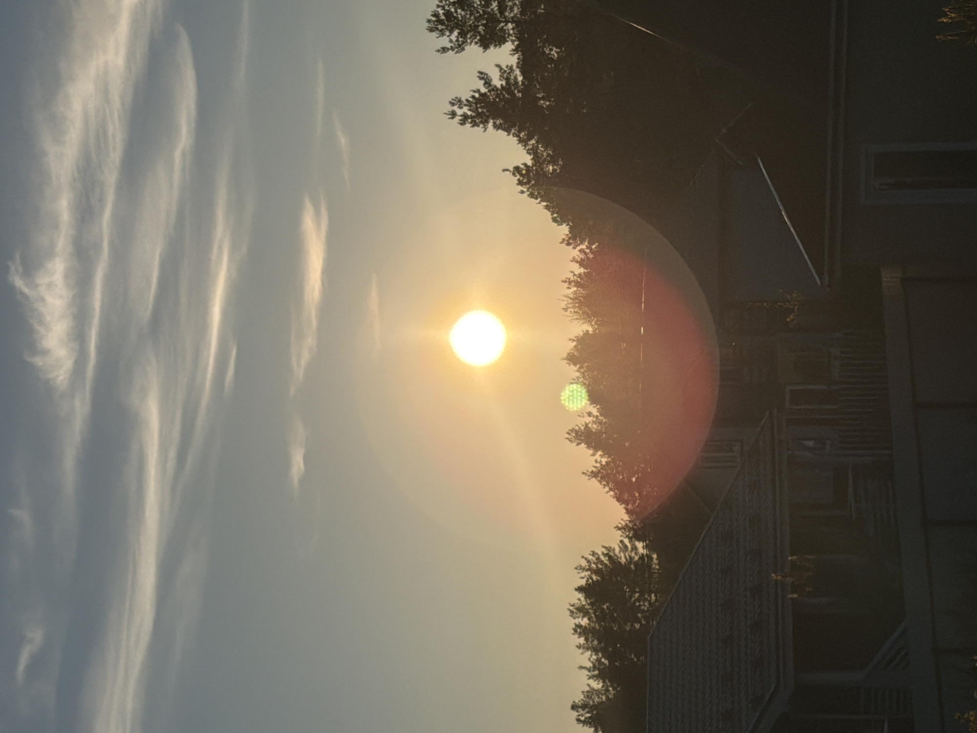 The image shows a sunset view in Ветеран Снт, Russia. The sun is the brightest object in the sky, with a lens flare effect creating a circular halo. Streaky cirrus clouds are visible in the left part of the sky. The silhouette of trees and a two-story house is visible on the right side of the image, obscuring part of the sun. A balcony with railings is visible on the house.