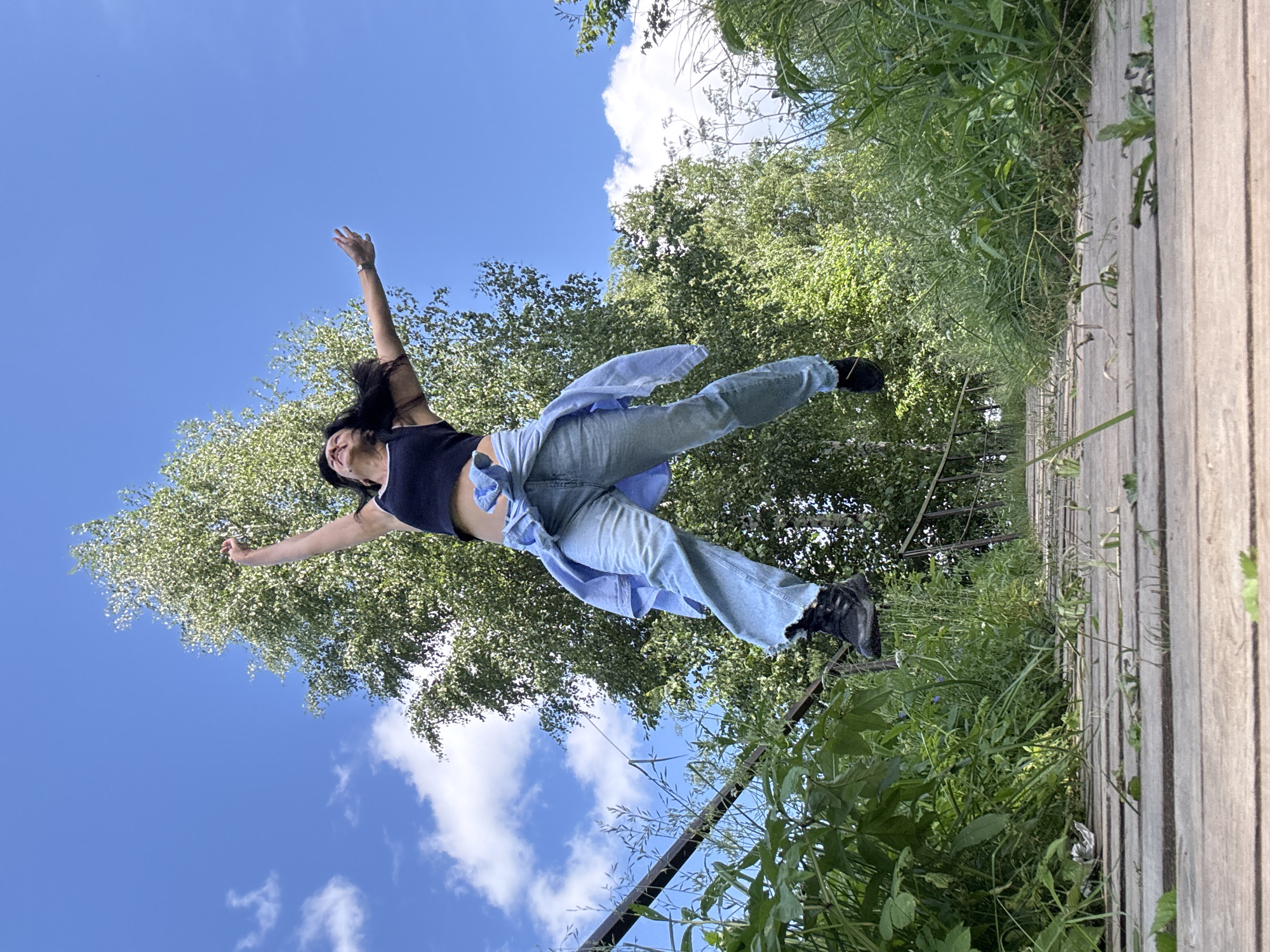 A woman is captured mid-air in a joyful jump on a wooden walkway. She is wearing a black crop top, light blue jeans, and black boots. A light blue shirt is tied around her waist. Her arms are outstretched, and her head is tilted back, looking towards the sky. Behind her, a tall tree with green leaves stands against a bright blue sky with scattered white clouds. The wooden walkway appears to be surrounded by lush green grass and vegetation, with a metal railing visible on the left side. The scene suggests an outdoor location, possibly a park or nature trail. It is not possible to determine the specific location within the city of Chaikovsky, Russia from the image alone.