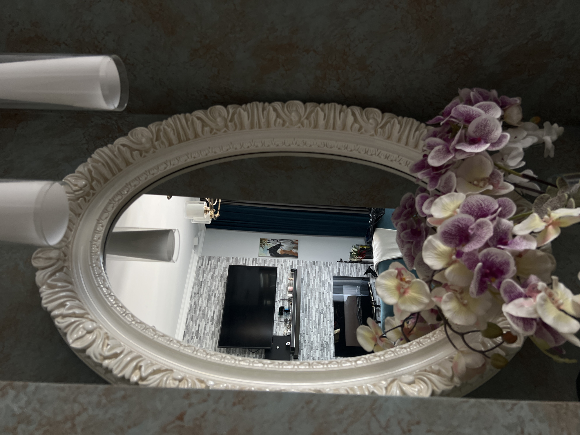 The image shows an oval, ornate mirror with a white frame, reflecting a living room. The reflected room features a modern fireplace with a television mounted above it, surrounded by a grey brick veneer wall. A dark shelf below the TV holds decorative items. To the right of the fireplace, a painting of horses is visible, and dark teal curtains hang to the right. In the foreground, a bouquet of artificial orchids in various shades of white and purple is placed in front of the mirror. The wall on the right side of the image has a textured, dark greenish-blue finish. Two cylindrical lampshades are visible hanging from the ceiling. No people are present in the scene. The image does not contain any information that would indicate its location in Rome, Italy.