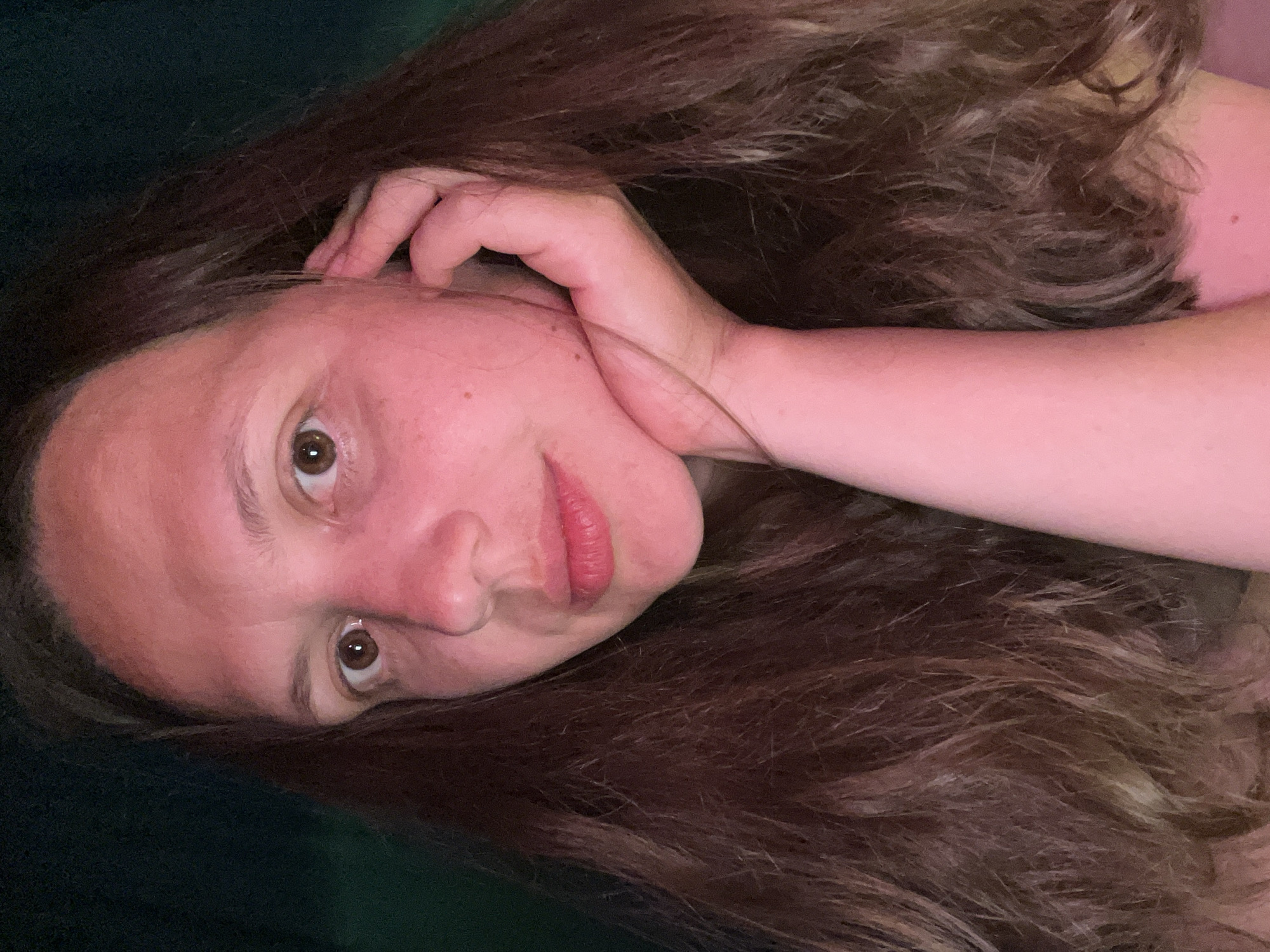 A woman with light skin and brown eyes is seen resting her head on her hand. She has long, brown hair with some lighter strands. Her lips are slightly parted and appear to have a reddish tint. She is lying on a dark green or black surface. The location is not discernible from the image. There is no information to suggest the picture was taken in Chaikovsky, Russia.