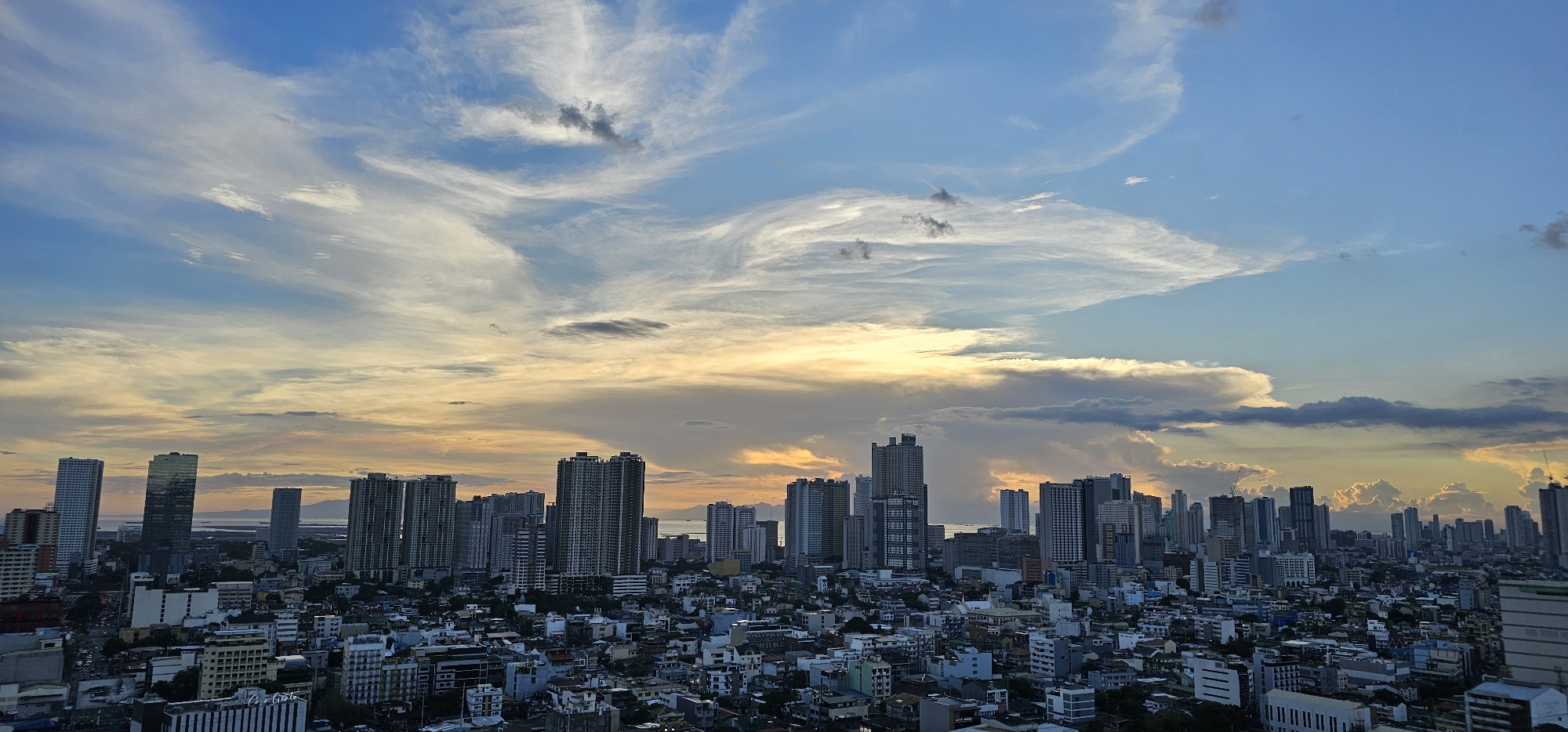 The image shows a panoramic view of the cityscape of Makati, Philippines, during what appears to be either sunrise or sunset. Tall buildings and skyscrapers dominate the horizon, indicative of a dense urban environment. The sky is a dramatic display of blues and oranges, with wispy clouds streaking across the expanse. The lower part of the image is filled with a dense collection of buildings, varying in height and architectural style, suggesting a well-developed metropolitan area. The lighting suggests ambient natural light, with shadows cast by the buildings. No people are discernibly visible in the image, nor are there any specific actions or interactions depicted. The scene is static and focuses on the architectural landscape and the atmospheric conditions of the sky.