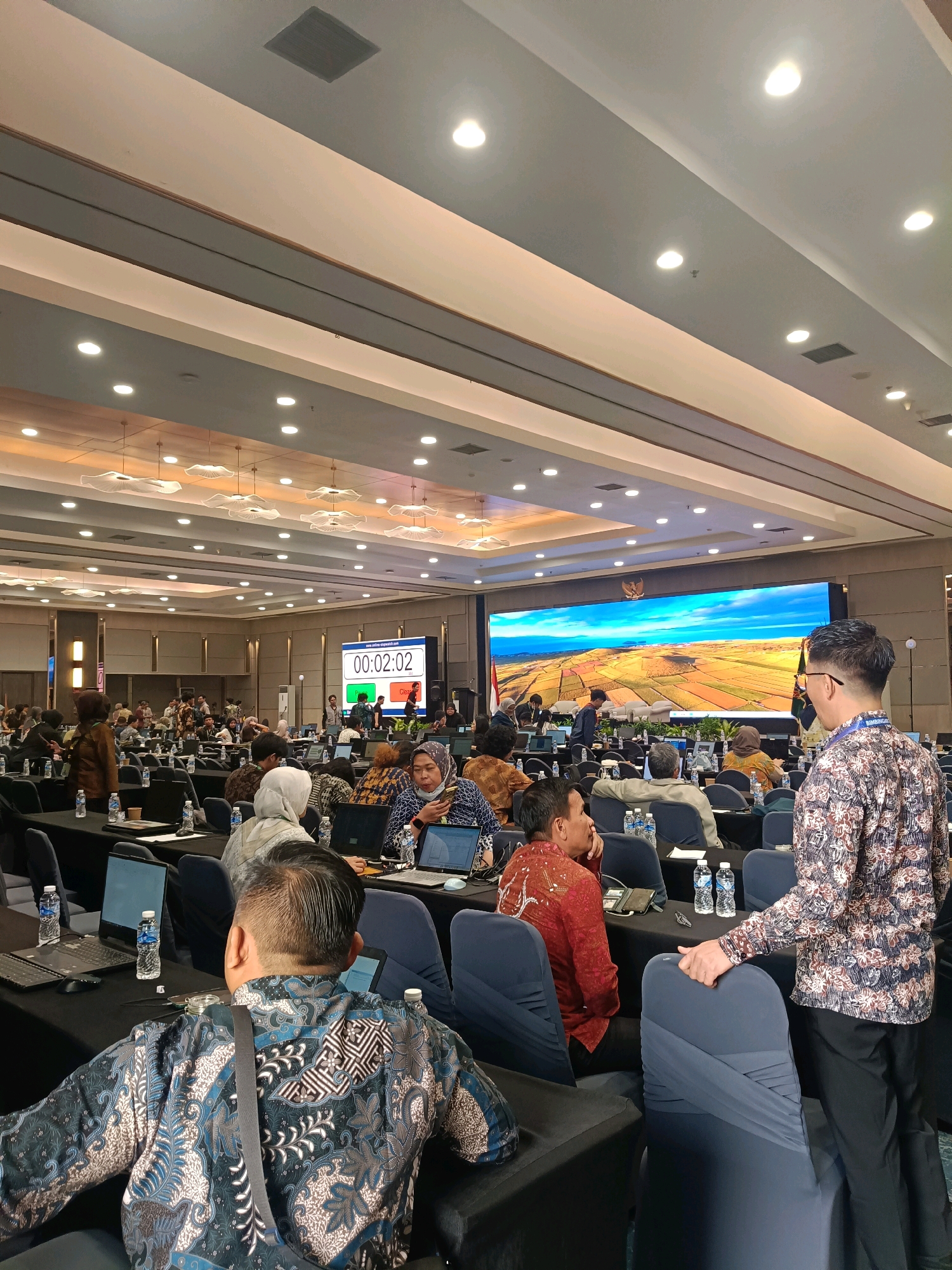 The image depicts a large conference room filled with attendees seated at long tables, facing a large screen at the front. The room is illuminated by overhead lighting and has a modern, spacious design. Several people are visible, some engaged with laptops, others looking towards the front, and one person in the foreground on the right is standing and addressing the audience. The screen displays a scenic landscape with fields and a body of water under a bright sky. A digital timer on a separate screen shows "00:02:02". The setting appears to be a formal event, possibly a conference or seminar, with water bottles and laptops arranged on the tables. The overall atmosphere is one of active participation and engagement.