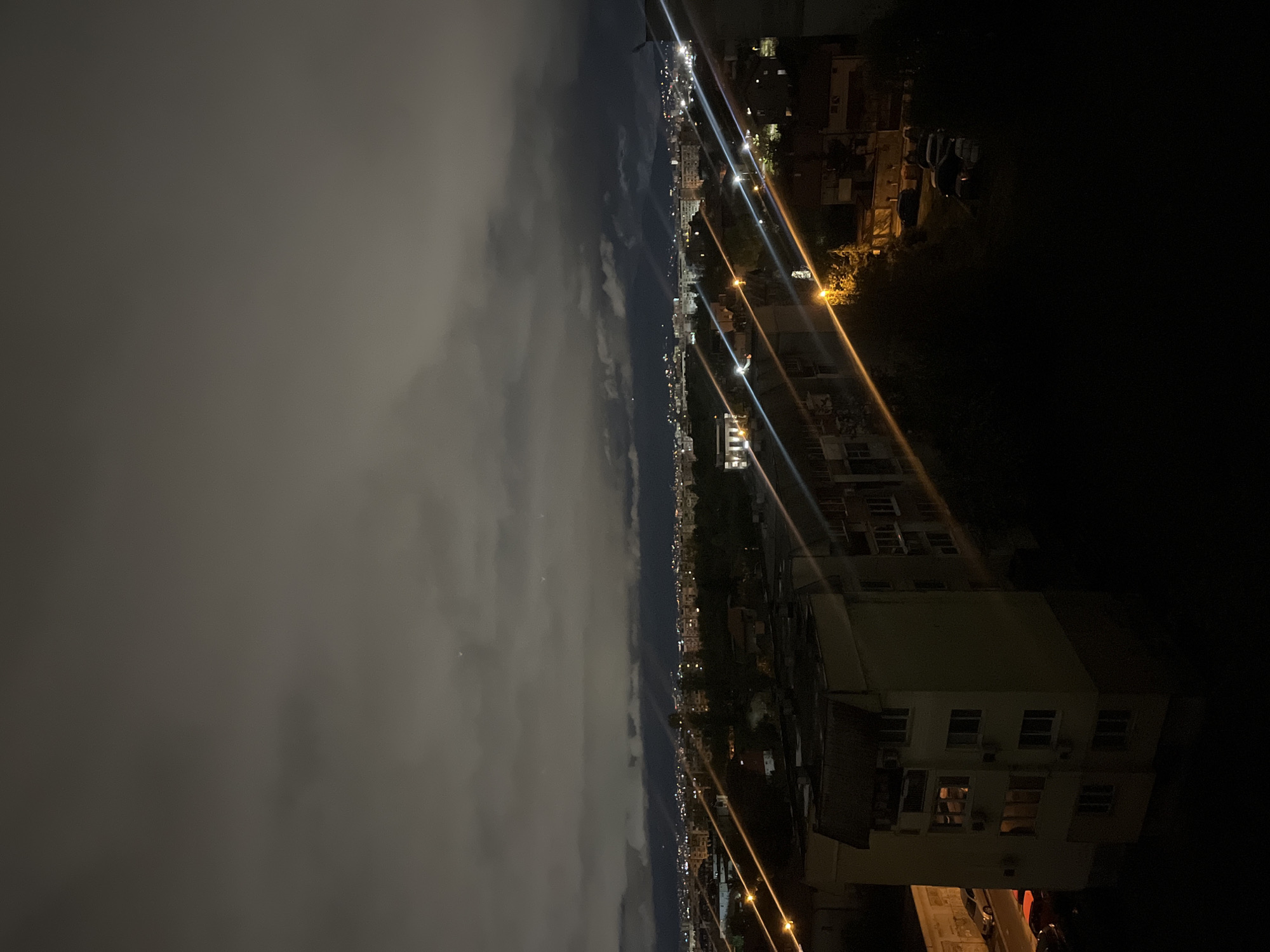 The image shows a nighttime cityscape, likely in Sofia, Bulgaria. The left side of the image is dominated by heavy cloud cover obscuring the horizon. To the right of the clouds, a dense urban area stretches into the distance, illuminated by streetlights and lights from buildings. The lighting creates linear streaks of illumination along streets. In the foreground on the right, a multi-story building is visible, with lit windows suggesting occupancy. The overall scene is dark, with the artificial lights providing the primary source of illumination.