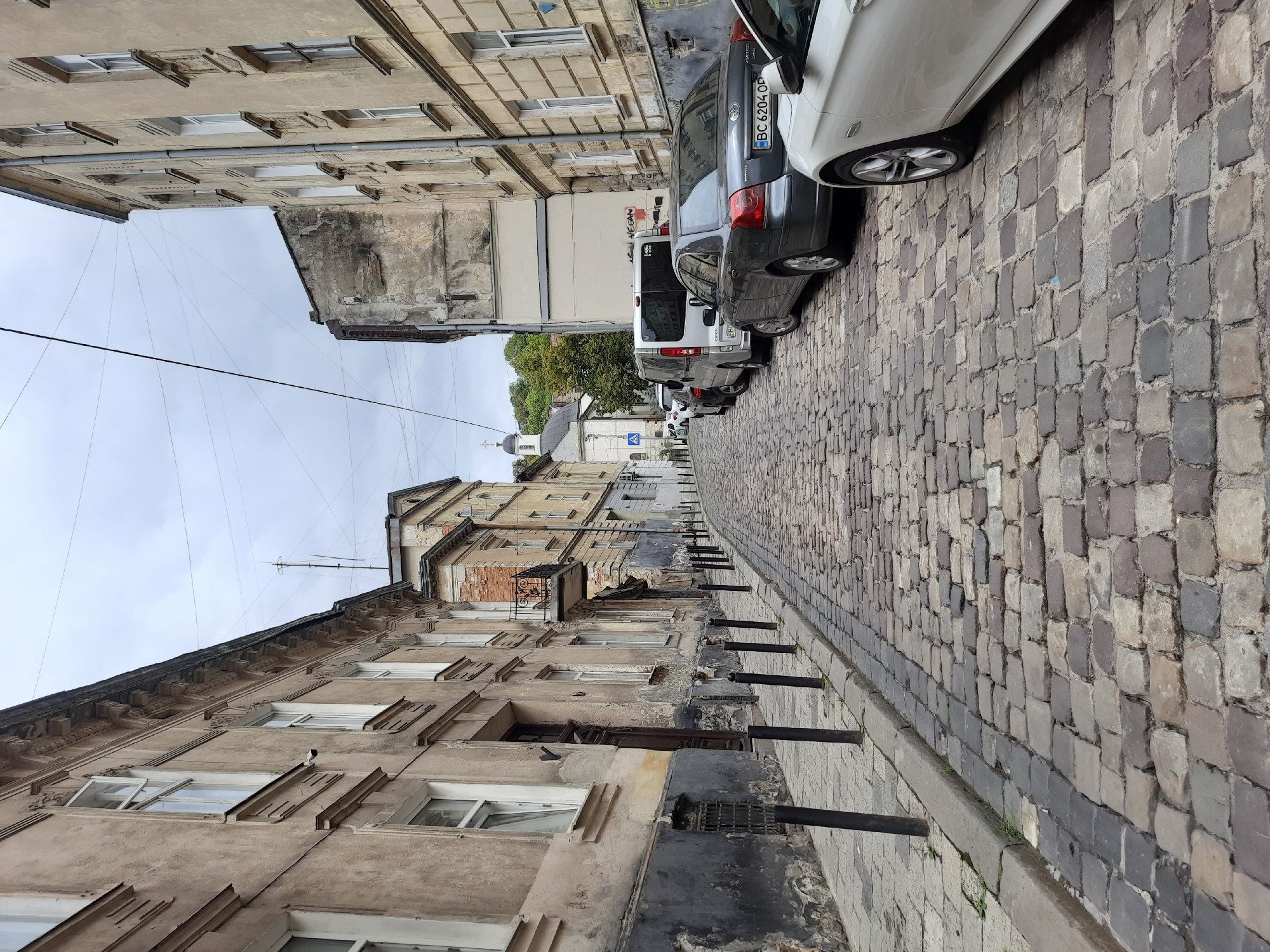 The image depicts a cobblestone street in Lviv, Ukraine, with buildings lining both sides. The street slopes upwards and curves to the right. On the left, older buildings with weathered facades and visible architectural details, such as cornices and window frames, are present. Some windows are open, and there is a dark wooden door. Black bollards are installed along the sidewalk on the left.
On the right side of the street, more buildings with similar architectural styles are visible, also showing signs of age. Several cars are parked along the right side of the street, including a white car in the foreground, a dark gray sedan, and a white van further up the street. In the background, a church with a prominent dome and cross can be seen, situated on a hill with trees. The sky is overcast, suggesting a cloudy day. The overall scene conveys a sense of historical urban architecture and a quiet street.