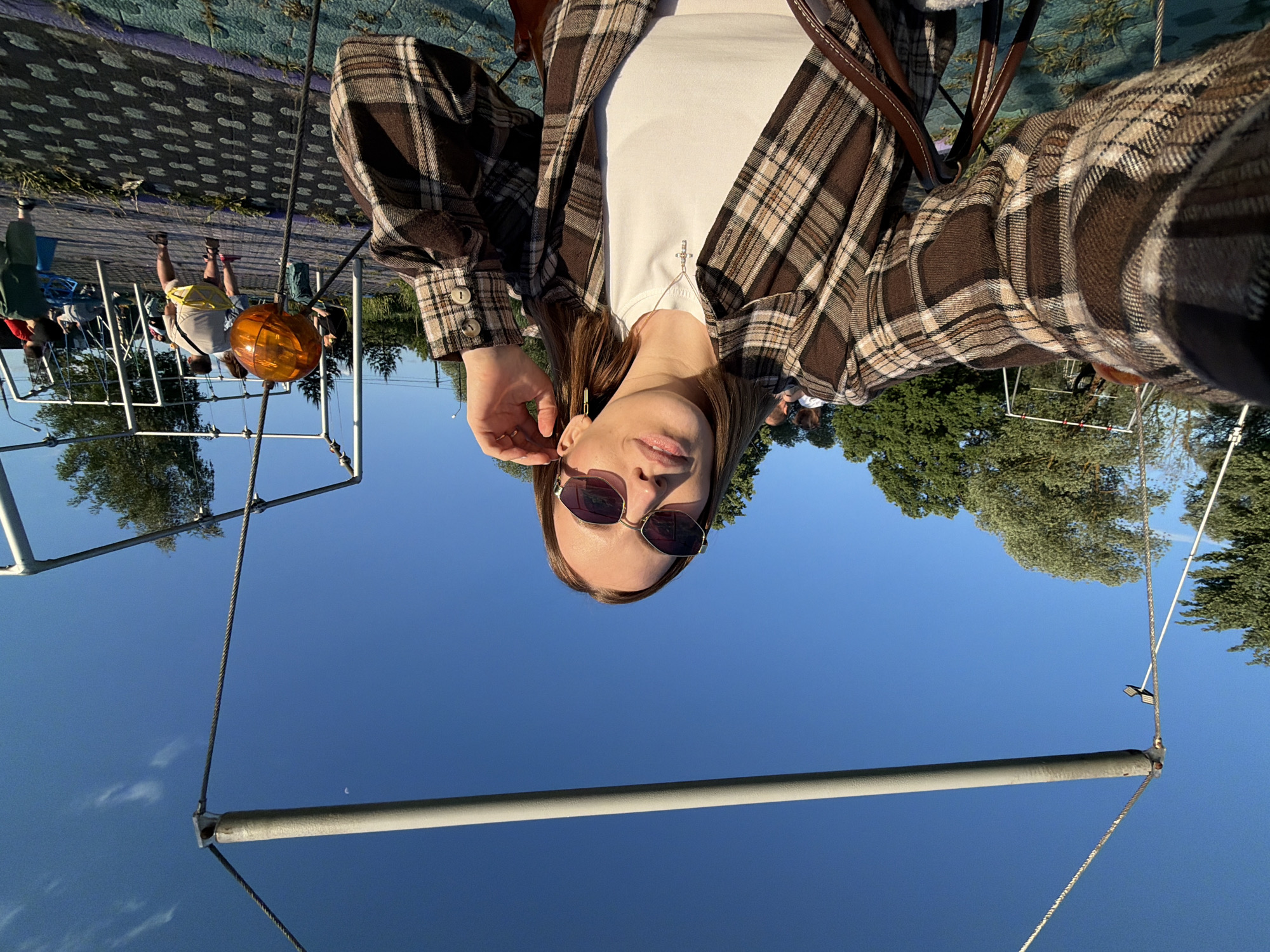 The image presents a woman in Sin'kovo, Russia, wearing sunglasses, a white t-shirt, and a brown plaid jacket. She has brown hair and is wearing a necklace. The background features a blue sky and trees. To her left, there is a playground structure with an orange ball hanging from it. People can be seen in the distance near the playground. The ground appears to be covered in a patterned surface.