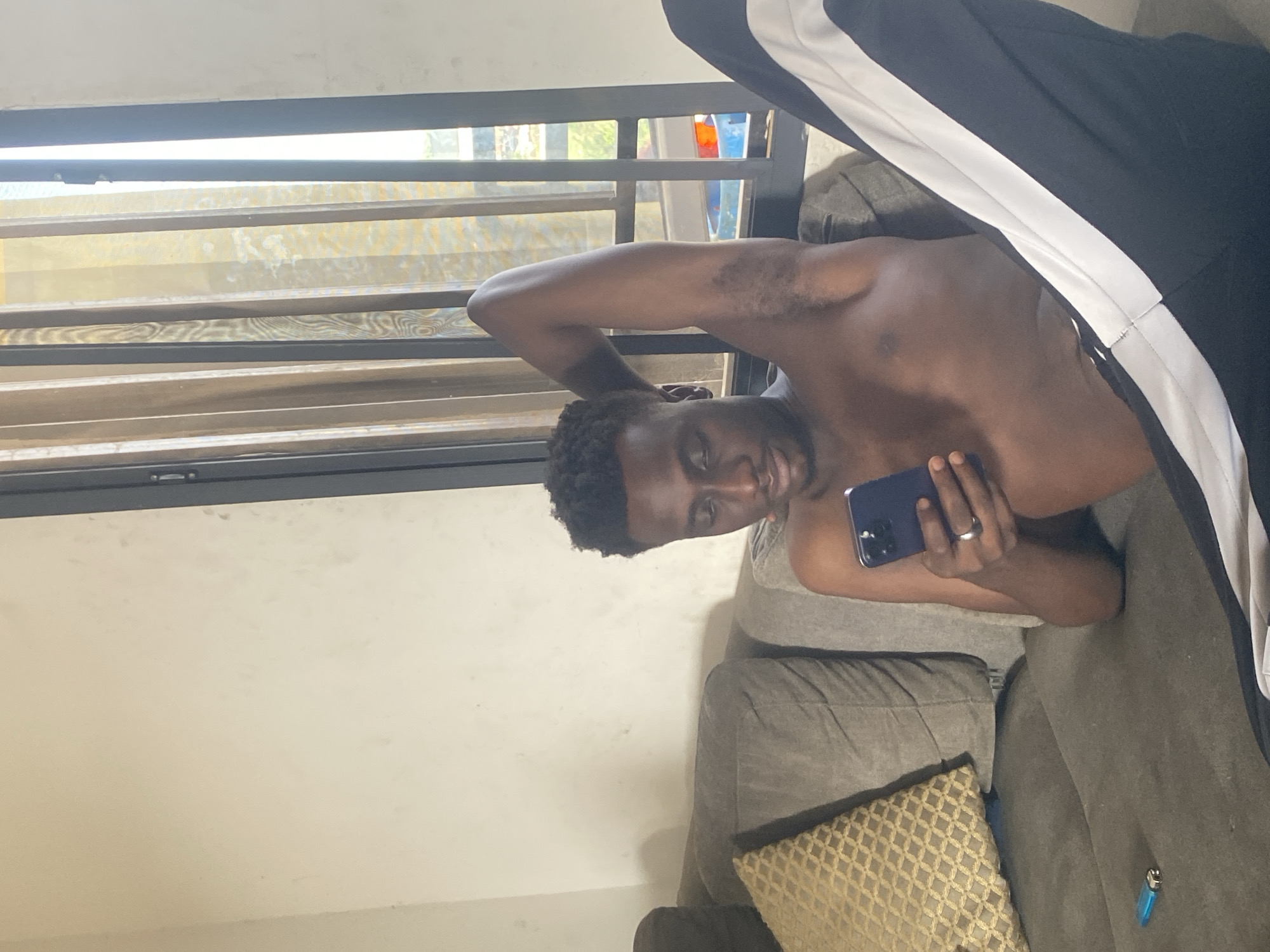 An indoor, horizontally oriented image captures a young Black man lying in a relaxed pose on a dark grey or muted brown upholstered couch. He is shirtless, displaying a muscular build, and wears black sweatpants with a wide white stripe down the side. The man has short, dark, curly hair and a light beard, and is looking towards the viewer with a soft smile.
His right arm is bent behind his head, while his left hand holds a dark-colored smartphone, possibly an iPhone, with a silver-toned ring on his left ring finger. Behind him, a large window fitted with horizontal slat blinds or louvers allows natural light to enter the room. Through the window, vague outlines of greenery and some colorful objects, resembling bottles, are visible outside.
To the left of the man, a plain, light-colored wall is visible. On the couch beside him, a decorative throw pillow with a golden-beige geometric pattern adds a touch of accent. A small, blue translucent lighter is also resting on the couch cushion near his hip. The setting appears to be a comfortable living room or lounge area, illuminated by daylight, suggesting a relaxed atmosphere in a residence in Abuja, Nigeria. There is no visible text in the image.
