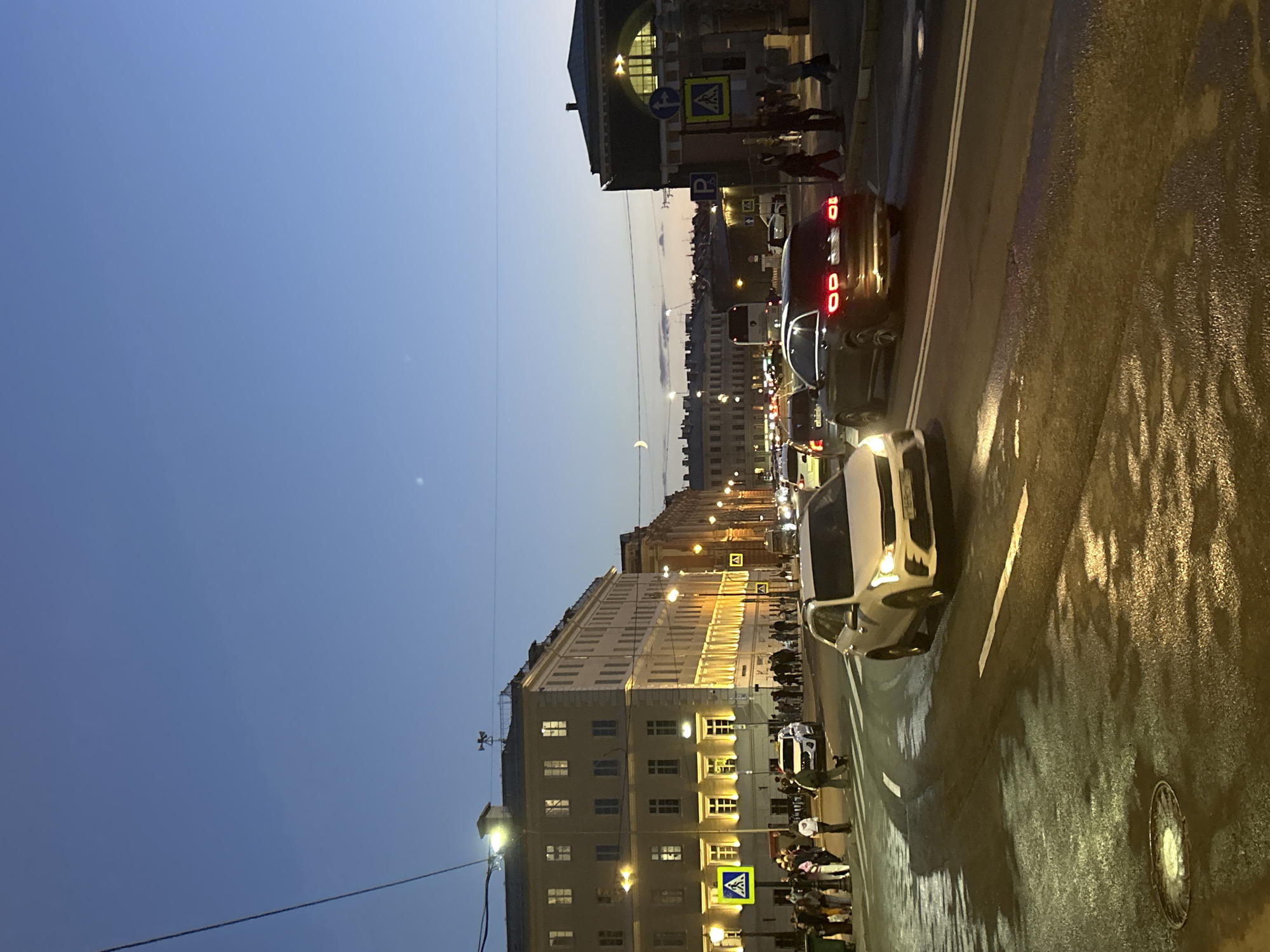 The image depicts a street scene at dusk in an urban environment. A light-colored SUV and a dark-colored sedan are driving on the wet asphalt road. Several pedestrians are visible on the sidewalk next to a large building with illuminated windows. Streetlights are on, casting a warm glow on the scene. In the background, more buildings line the street, and the sky is a deep blue, indicating the transition from day to night. There are also traffic signs and overhead wires present. The overall impression is of a busy city street during the evening hours.