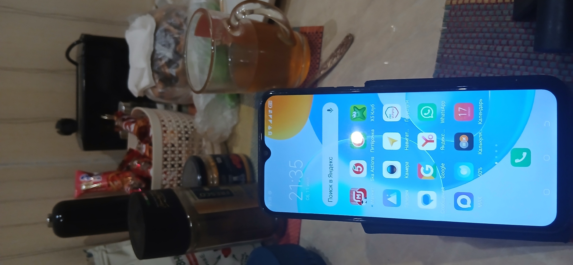 The image shows a smartphone displaying the lock screen with the time 21:35 and the date Сб, 11 июл. The phone is placed on a table with various objects, including a glass of tea, jars, and what appears to be food items in a basket. The background suggests an indoor setting. No people are visible in the image. The location in Verkhny Uslon, Russia, is an assumption based on the provided text and cannot be confirmed from the image itself.
