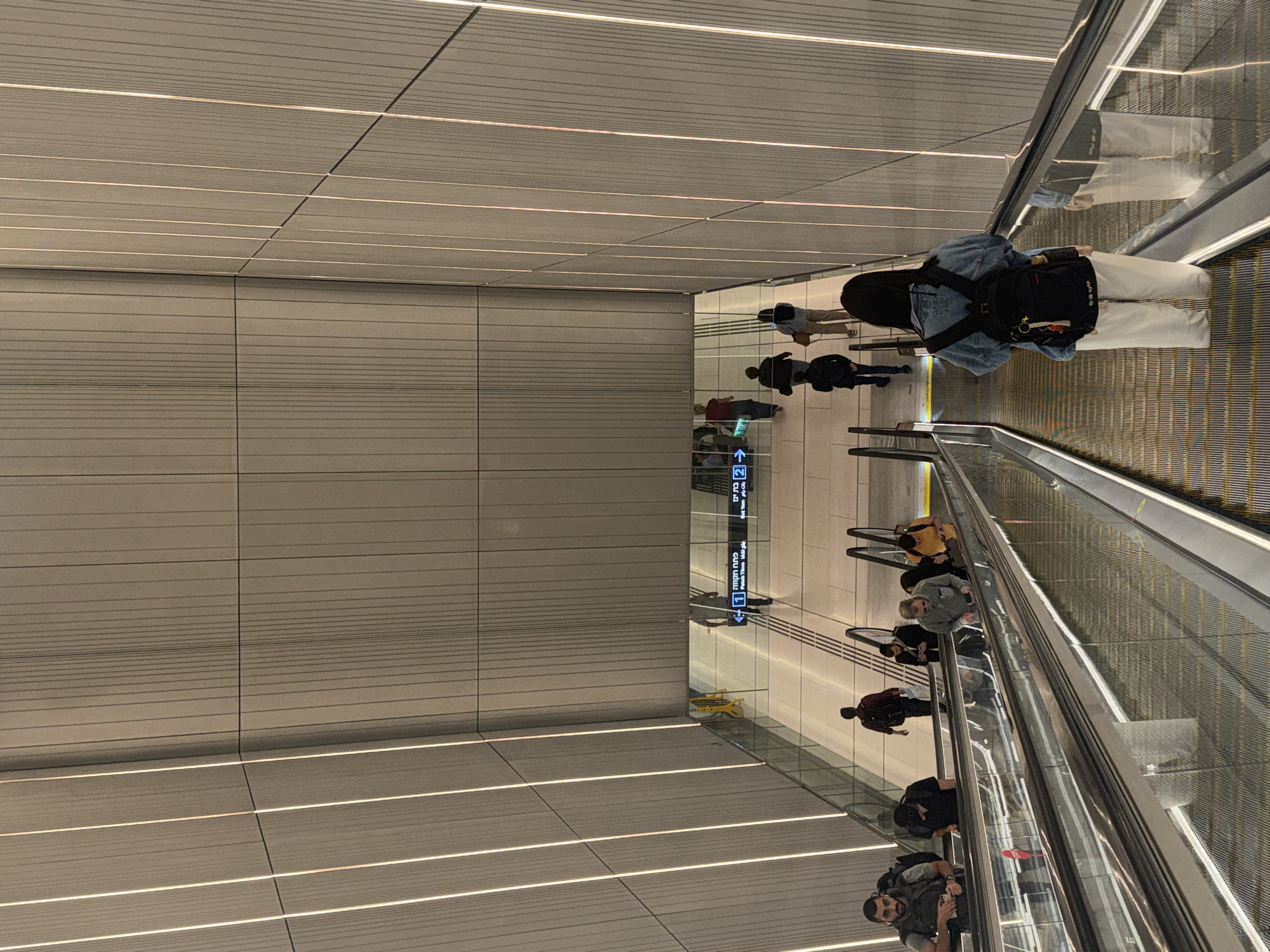 The image shows an interior scene in Ramat Gan, Israel. Several people are on an escalator. Some individuals are walking on the flat floor. A sign with Hebrew script is visible. The escalator has metallic guardrails and yellow stripes. The walls and ceiling consist of rectangular tiles. Overhead lighting is present.