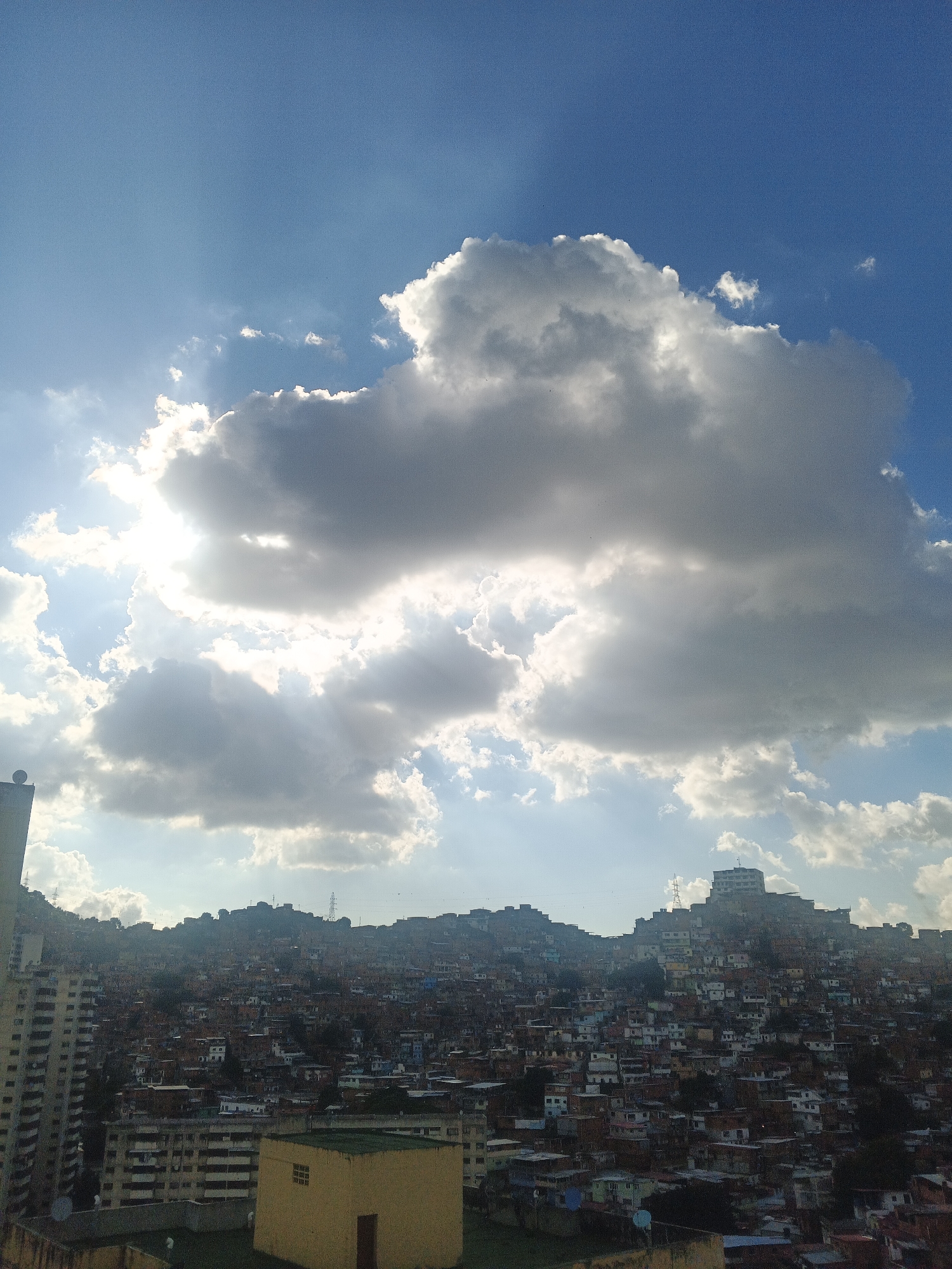 This image captures a panoramic view of a densely populated urban hillside in Caracas, Venezuela, likely a barrio or favela, set against a dramatic sky filled with large, billowing clouds. The sun is partially obscured by a prominent cloud formation, casting rays of light that illuminate parts of the sky and create dramatic shadows across the landscape.
The hillside is covered with an intricate patchwork of buildings, primarily single-story and multi-story structures, many with brightly colored roofs and walls. The architecture appears to be a mix of informal construction and more established housing. The density of the settlement is striking, with buildings tightly packed together, climbing the natural contours of the hill.
In the foreground, to the left, a tall, modern apartment building stands out, its facade a grid of windows and balconies. To the right and center foreground, a low-rise, light yellow building with a single window is visible. Further back, the hillside is dominated by the informal settlements, with clusters of houses creating a textured, layered effect. In the distance, the topography suggests a mountainous or hilly terrain surrounding the city.
The sky is a vibrant blue, contrasting with the gray and white clouds. The lighting suggests it might be late afternoon or early morning, given the dramatic interplay of light and shadow. There are no visible people or active interactions depicted in the scene. The overall impression is one of a vast, sprawling urban environment integrated with the natural landscape, under a dynamic sky.