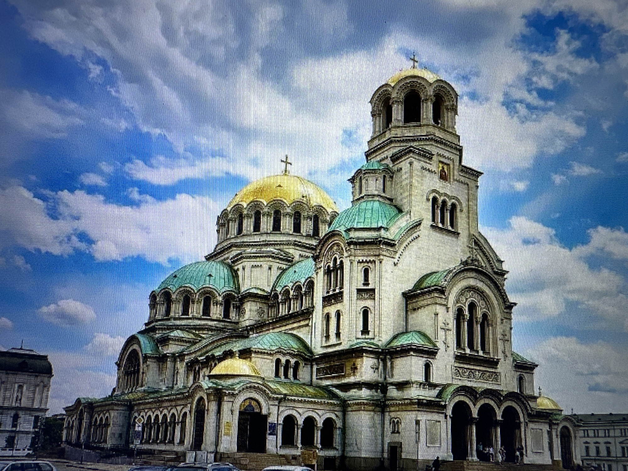 The image depicts the Alexander Nevsky Cathedral, a prominent landmark in Sofia, Bulgaria, not Kostenets. The cathedral is a massive Orthodox church with a striking architectural style characterized by multiple domes, predominantly green and gold, and intricate stonework. A large golden dome with a cross is visible in the background, while several smaller green domes adorn the main structure. The facade features arched windows, columns, and decorative carvings. Several people are seen near the entrance on the steps, appearing as small figures against the grand scale of the building. The sky above is a vibrant blue, filled with scattered white clouds. In the foreground, a street level view shows parts of other buildings and what appear to be vehicles, suggesting an urban setting. The lighting suggests it is daytime, with the sun illuminating the facade of the cathedral.
