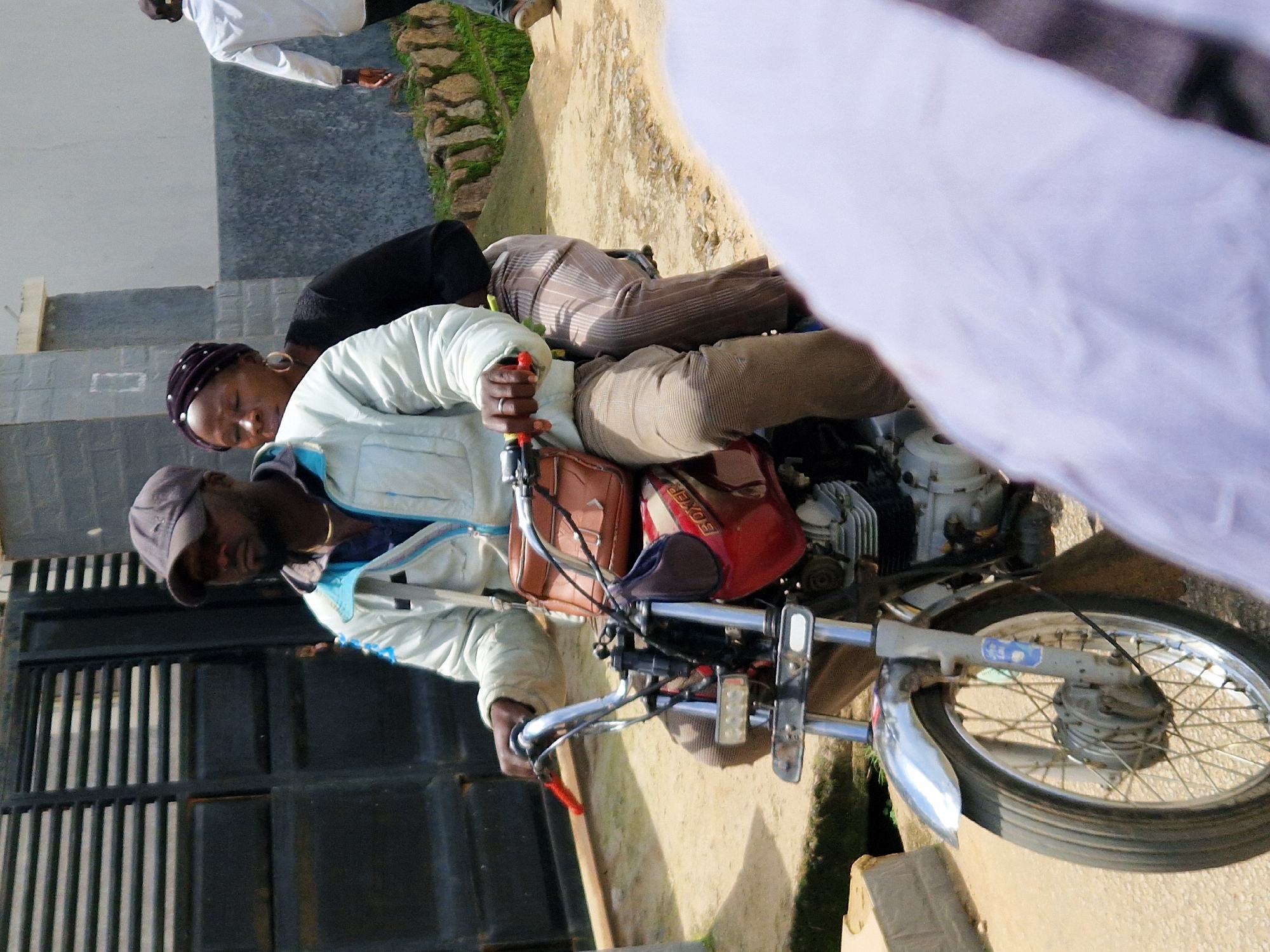 A man wearing a dark cap and a light-colored jacket is riding a motorcycle, with a woman wearing a dark headscarf and a black top as a passenger. The motorcycle is red and appears to be a Boxer model. The man has a brown satchel on his left shoulder. They are on a dirt road in front of a dark metal gate and a building. Another person in a white shirt is walking away in the background. The scene appears to be in a rural or semi-urban area.