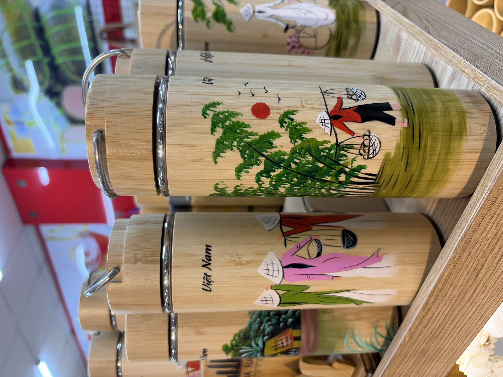 A group of cylindrical bamboo thermoses with decorative paintings are displayed on a wooden shelf. The paintings depict traditional Vietnamese scenes. One thermos features the text "Việt Nam" and shows three figures in conical hats carrying baskets. Another thermos shows a single figure in a conical hat carrying baskets, with a red sun and birds in the background. A third thermos displays a figure in white with a bicycle and pink flowers. The thermoses have metal handles and caps. The background is blurred, showing shelves with other merchandise, suggesting a retail environment. The specific location in Đà Nẵng, Vietnam cannot be determined from the image.