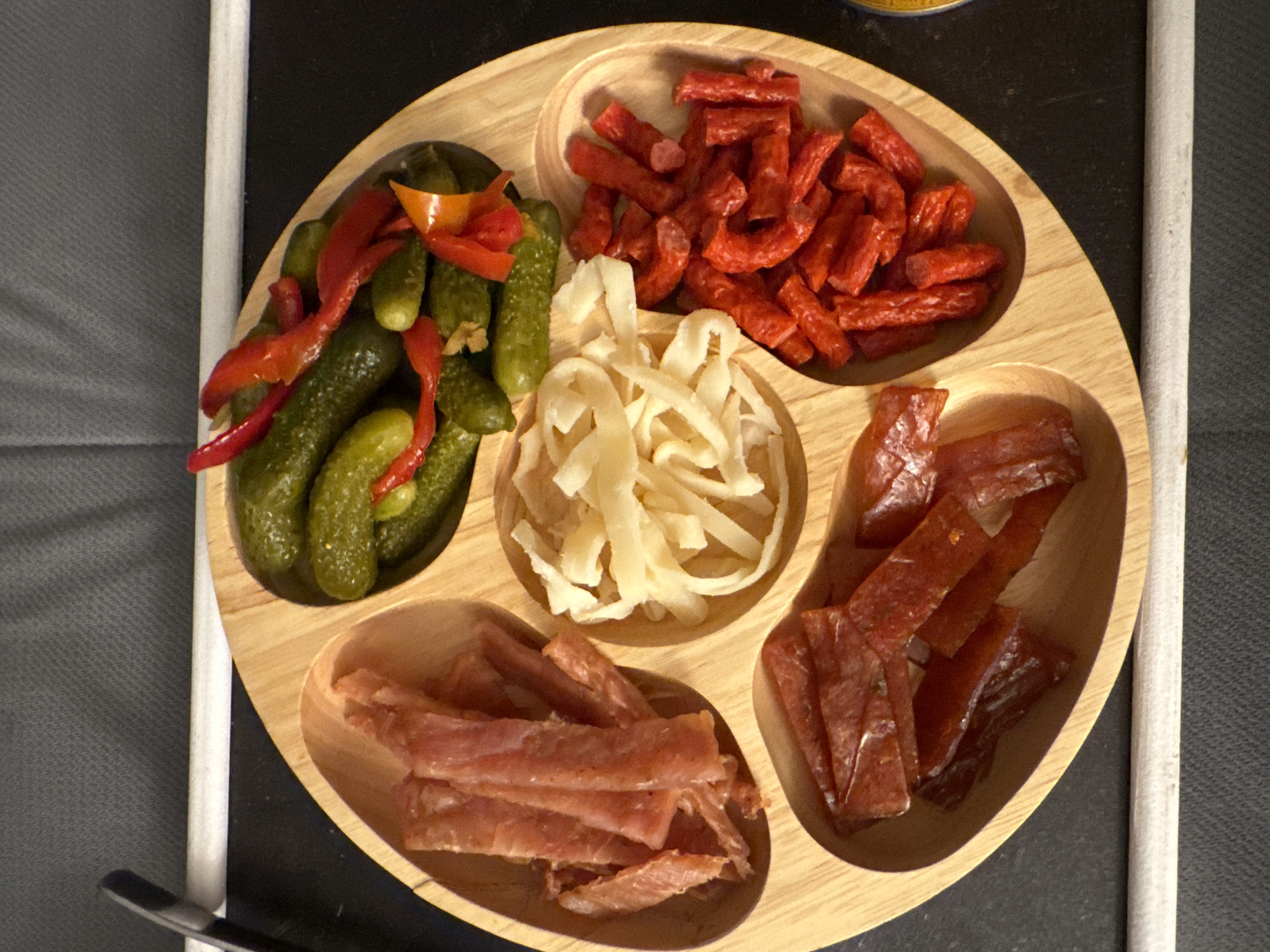 A wooden tray with five sections contains different food items. The top left section holds pickles and red pepper strips. To the right of that, a section contains cut-up sausages. Below the sausages, in the center, is a pile of white stringy cheese. To the right of the cheese, a section holds pieces of jerky. The bottom left section contains strips of light-colored meat. The tray sits on a dark surface. The location and time are unknown.
