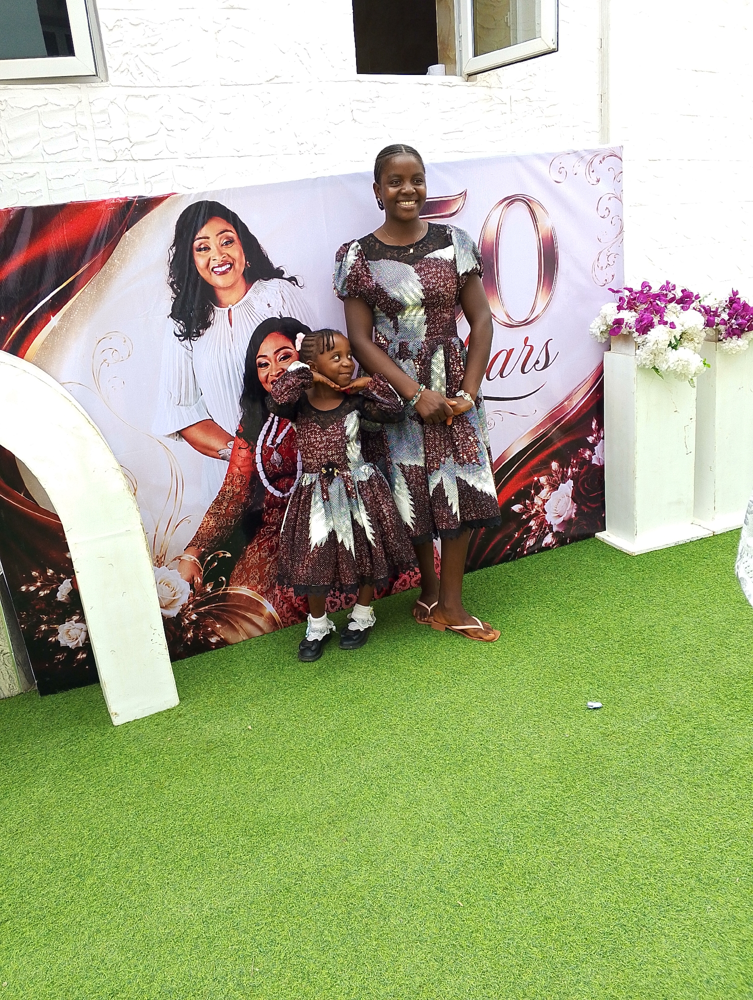 A woman and a young girl stand on artificial green turf in front of a large backdrop, likely at a celebration. The backdrop features a portrait of two women and the words "50 Years" in a stylized font. The woman is wearing a knee-length dress with a dark, patterned fabric and brown sandals. The young girl is also wearing a matching dress with dark shoes and white socks. She has her hands placed under her chin and is looking to the side. A white arched structure is visible to the left of the backdrop, and a flower arrangement stands to the right. The background suggests an outdoor setting with a building visible in the upper portion of the frame. This scene appears to have taken place in Bar Jirgi Summa, Nigeria.