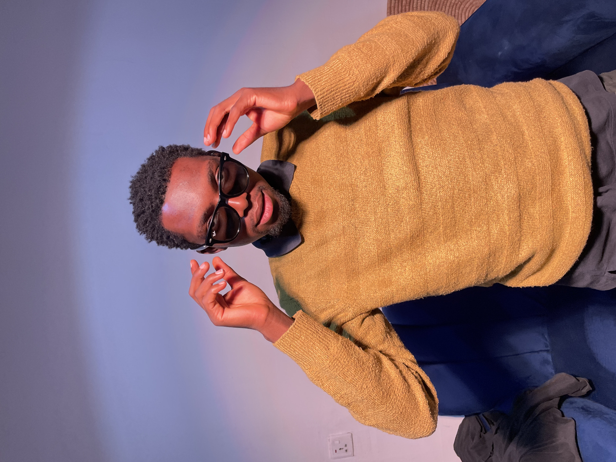 A man with dark skin, short curly hair, and a beard sits on a blue sofa. He is wearing a tan sweater, dark pants, and black-rimmed sunglasses. His hands are raised, with fingers slightly spread. A black collared shirt peeks out from beneath the sweater. The background is a light gradient from blue to white. A white electrical outlet is visible on the wall near the bottom of the frame. The scene is indoors. The location of the photo is not discernible from the provided image. Therefore, it is not possible to confirm it is in Barakin Akawo, Nigeria.