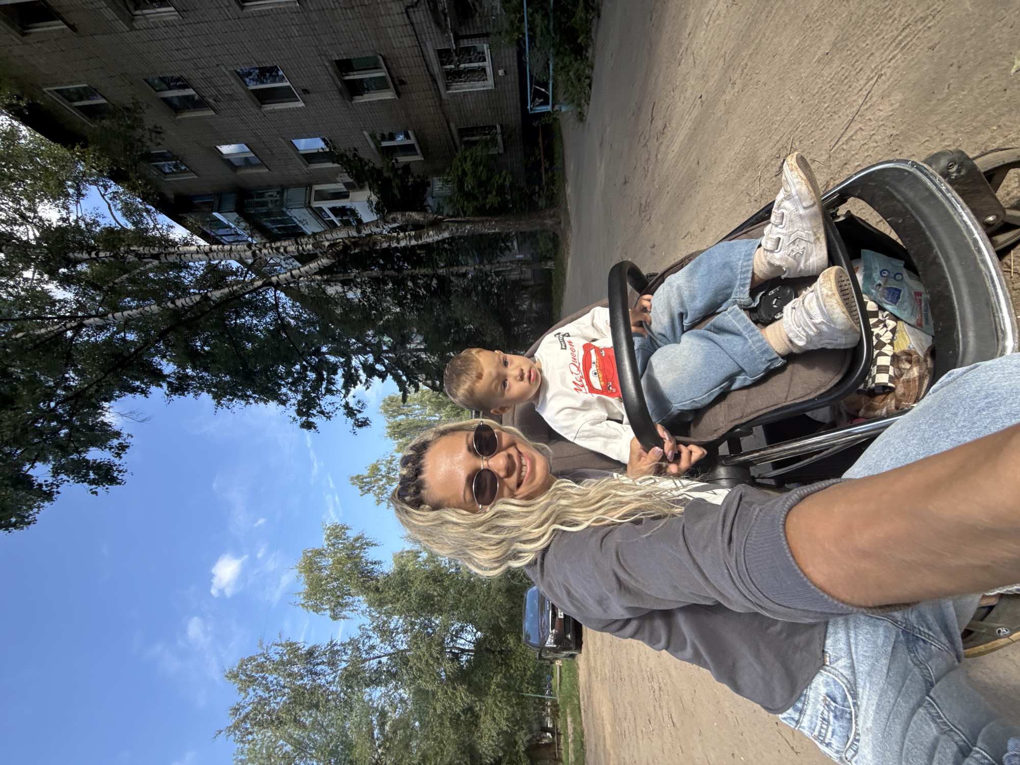 In this image, a woman with blonde, wavy hair and sunglasses is taking a selfie while pushing a stroller. She is smiling at the camera, conveying a happy emotion. Seated in the stroller is a young boy wearing a white long-sleeved shirt with a red Lightning McQueen graphic and blue jeans. He appears to be looking away from the camera. The woman is wearing a grey sweatshirt and blue jeans.
The scene is outdoors, with a paved pathway in the foreground. To the left, a multi-story brick apartment building is visible, with trees and lush green foliage surrounding it. The sky is a clear blue with a few wispy clouds, suggesting a pleasant day. The time of day appears to be daytime, possibly late morning or early afternoon, judging by the lighting.
The overall setting indicates a residential area, likely a courtyard or a path in a park adjacent to apartment buildings. The image is oriented such that the sky is at the top and the ground is at the bottom, but the subjects are angled. The stroller appears to be a common baby stroller with a black frame and fabric seat. A small blue packet with some writing is visible inside the stroller. The woman's hand is visible holding onto the stroller's handle, and the boy's hands are also visible within the stroller. The context of "Rusanovo, Russia" places the scene in a specific geographical area within Russia.