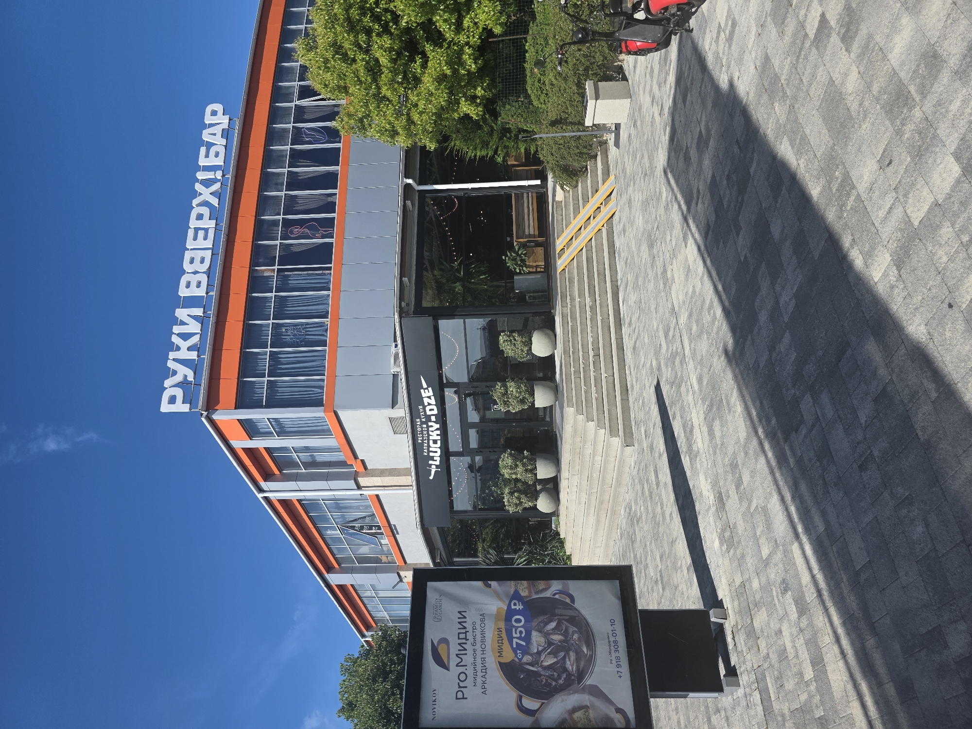 This full-shot image captures a commercial building and its immediate surroundings on a clear, sunny day in Sochi, Russia, identified by text as being near the "Mandarin Entertainment Complex."
The main subject is a two-story building featuring a gray facade with prominent orange bands and window frames on the upper level. The roof displays a large white sign reading "РУКИ ВВЕРХ! БАР" (Hands Up! Bar). The upper floor windows are largely reflective, some showing interior curtains, and at least two distinct neon-style signs (one resembling a flamingo, another a lightning bolt shape) are visible through the glass.
The ground floor houses the "LUCKY-DZE" restaurant, identified as a "РЕСТОРАН КАВКАЗСКОЙ КУХНИ" (Restaurant of Caucasian Cuisine). Its entrance is characterized by a black-framed glass facade, accessible via a series of six gray concrete steps. A pair of yellow, metal wheelchair ramps are positioned on the right side of these steps. The entrance area is adorned with multiple large potted plants, and string lights are visible within the interior along with wooden bench seating.
In the foreground, a paved plaza composed of alternating light and dark gray rectangular pavers extends across the scene. On the left, a black advertising stand prominently displays an advertisement for "Pro.Мидии" (Pro.Mussels) by "NOVINIKOV" and "FAMILY GARDEN," offering mussels starting from 750 rubles. The ad also includes a Russian phone number (+7 918 308-01-10) and the reference to "РК «Мандарин»" (Mandarin Entertainment Complex). Long, dark shadows from the advertising stand and potentially other objects are cast across the paved ground, indicative of direct sunlight.
Green foliage, including trees and bushes, is visible on both sides of the building. On the far right, a portion of a red scooter or similar two-wheeled vehicle is visible alongside a gray concrete trash receptacle. The sky above is a clear, vibrant blue with minimal wispy cloud cover. No individuals are present in the frame.
