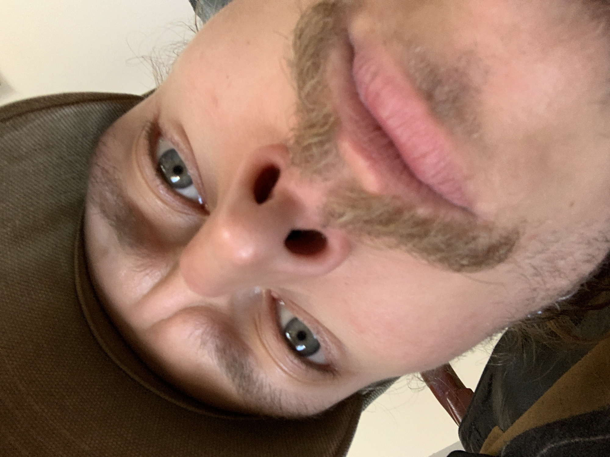 The image shows a close-up, distorted, and downward-angled view of a person's face. The person has light skin, a mustache, and gray eyes. Part of a brown hat is visible on the left side of the frame. The background is blurry and indistinct. There is no discernible information to indicate that this image was taken in Tel-Aviv, Israel.