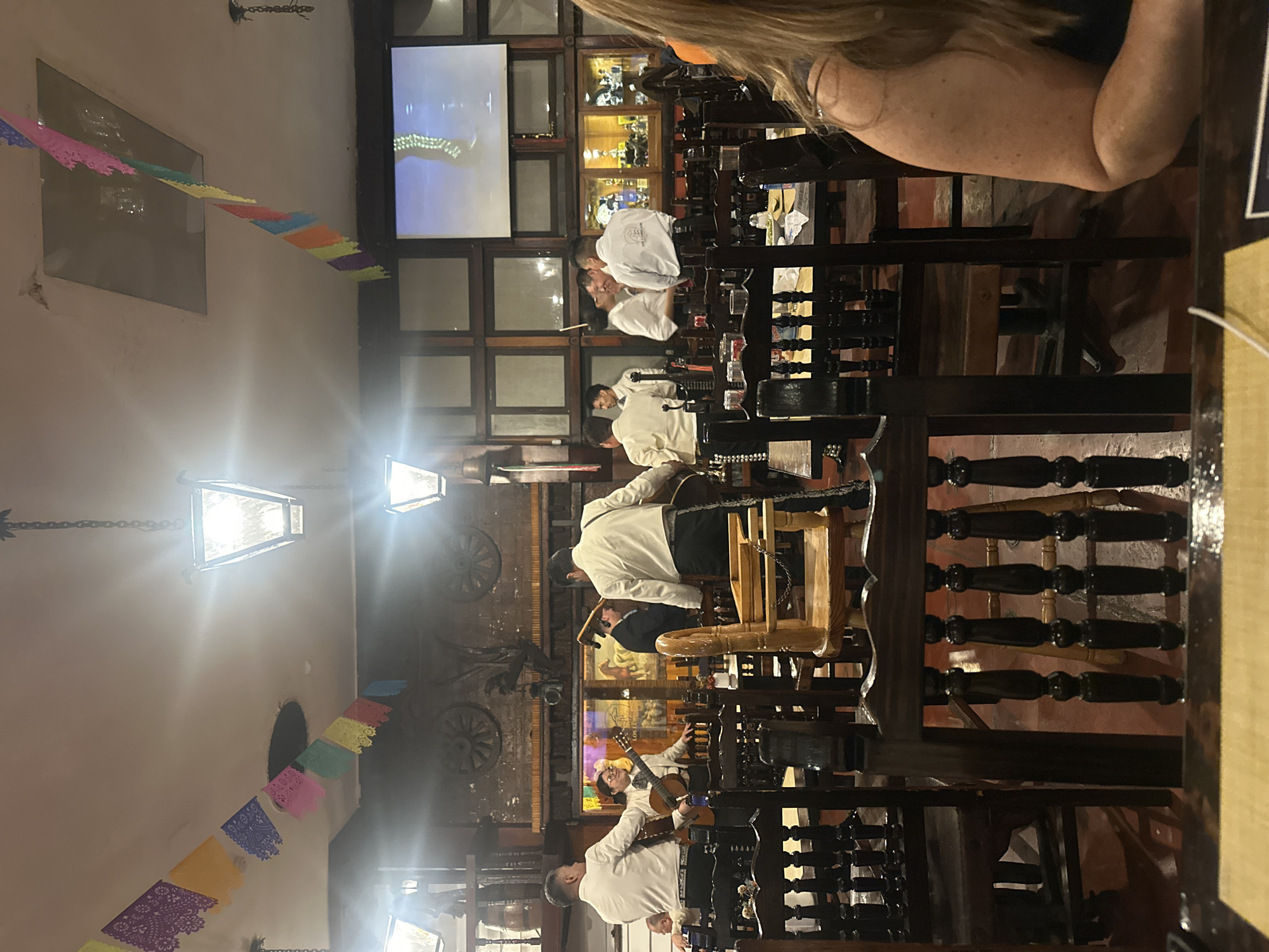 The image shows a group of mariachi musicians performing in what appears to be a restaurant in Mexico City, Mexico. The band members are dressed in traditional white mariachi attire, playing instruments such as guitars and a violin. The restaurant has a warm, rustic atmosphere, with dark wood furniture and decorations. Colorful papel picado banners are strung across the ceiling. A television screen is visible in the background. Diners are seated at tables, with one person’s shoulder and arm visible in the foreground.