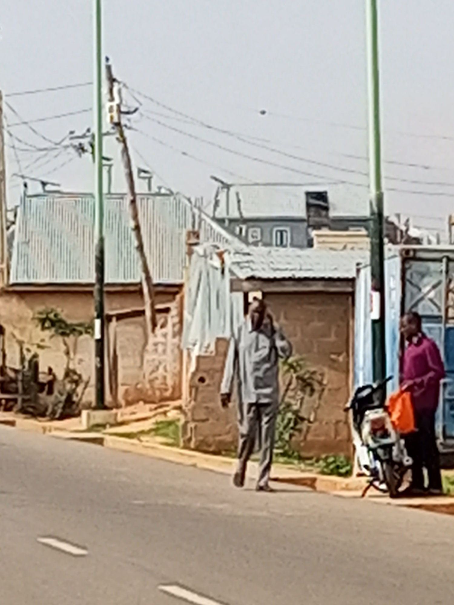 The image depicts a street scene in Gombe, Nigeria. In the foreground, a paved road with white lane markings is visible. In the mid-ground, two men are present. One man, on the left, is wearing a light-colored collared shirt and trousers, and appears to be speaking on a mobile phone. He is walking towards the right of the frame. To the right of this man, a motorcycle is parked. Standing near the motorcycle is a second man, wearing a dark-colored shirt and trousers, holding an orange bag. In the background, there are buildings with corrugated metal roofs and brick walls, as well as utility poles and overhead wires. The overall impression is of an outdoor, daytime urban environment.