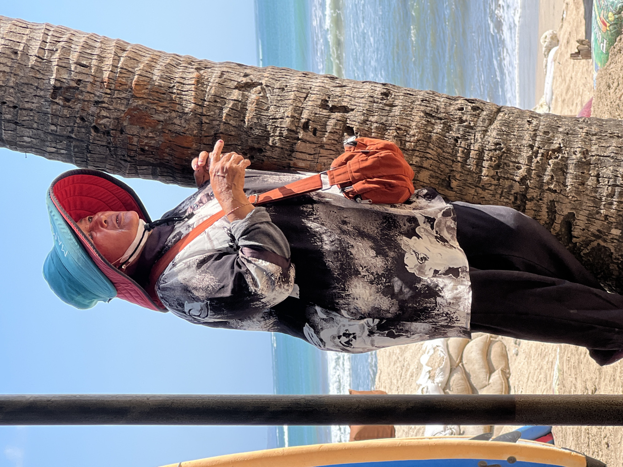 A person stands on a sandy beach in Kuta, Indonesia, leaning against a palm tree. They are wearing a black and white patterned shirt, dark pants, and a wide-brimmed hat with a turquoise top and red underside. An orange shoulder bag is slung across their chest. The person is looking towards the right side of the frame, with their right index finger extended as if pointing or speaking. In the background, the turquoise ocean meets the horizon under a clear blue sky. To the left, a portion of a surfboard and a dark pole are visible. Sandbags are stacked in the mid-ground behind the person.