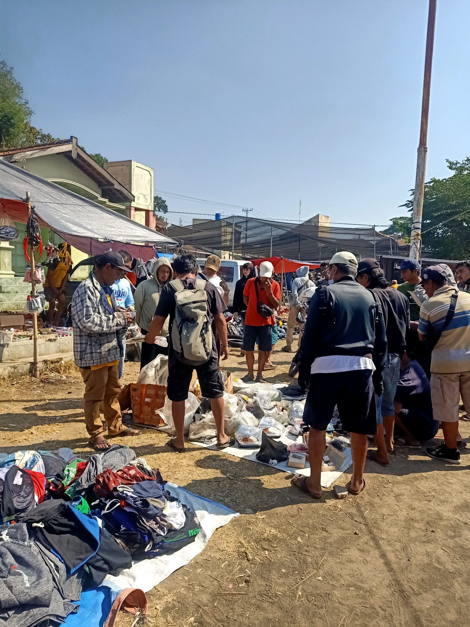 The image depicts an outdoor marketplace scene in Jatinom, Indonesia. A group of men are gathered around stalls, with some looking at merchandise laid out on the ground and others engaged in conversation. Clothing items, including shirts and shorts, are spread on a white tarp in the foreground, suggesting a sale or exchange. Various items, possibly related to electronics or other goods, are visible in plastic packaging behind the people. The background shows structures resembling shops or market stalls with tents, along with trees and a clear blue sky. A tall, weathered pole stands prominently on the right side of the image. The ground is dirt and dry vegetation.