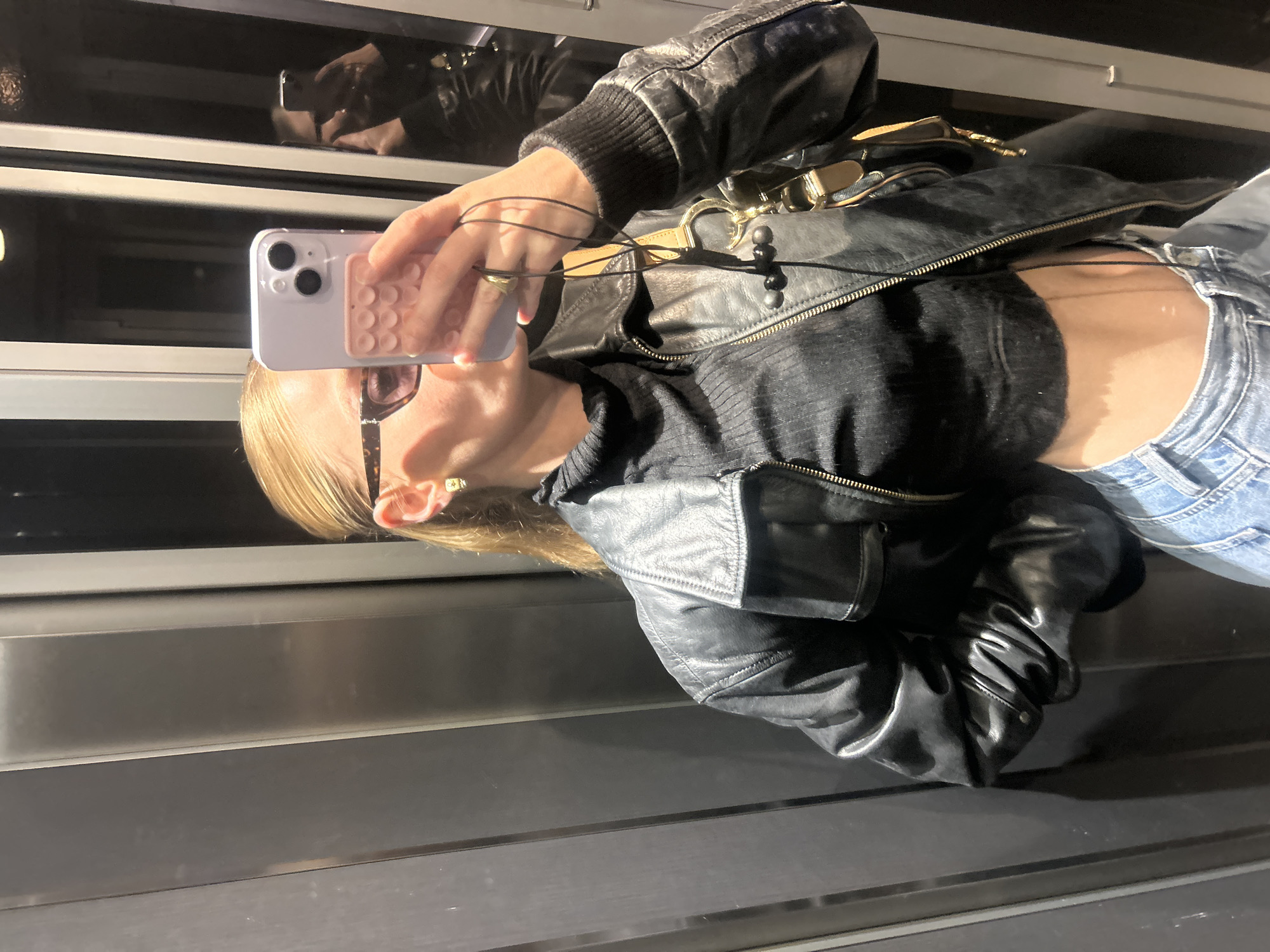 A selfie shows a person with blonde hair wearing a black leather jacket, black turtleneck, and light blue jeans. The person is holding a light purple smartphone in their right hand, and they are wearing glasses. There is a reflection in the glass behind them of the person taking the picture wearing a jacket. The background appears to be an elevator or some sort of transportation vehicle. The location is unidentified.
