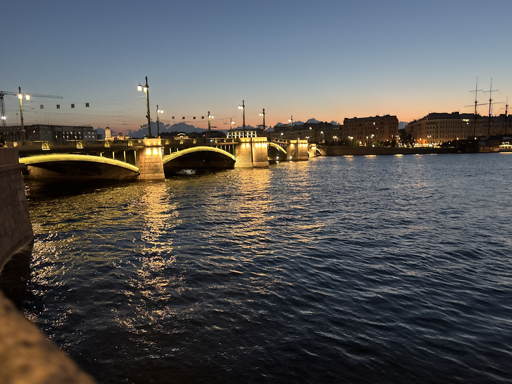 This is a nighttime photograph of a bridge and waterfront in Saint Petersburg, Russia, likely taken during the twilight hours. The bridge, illuminated by warm yellow lights, spans a dark body of water, likely a river or canal, reflecting the light. The water's surface is rippled. In the background, along the waterfront, are buildings with illuminated windows, suggesting a cityscape. To the right, a tall-masted ship is docked. The sky above is a gradient of deep blue and orange, indicating the transition from day to night. The image appears to be taken from a slightly elevated position, looking across the water towards the bridge and city. There are no visible people or specific actions depicted. The overall scene conveys a serene and picturesque urban landscape at dusk.