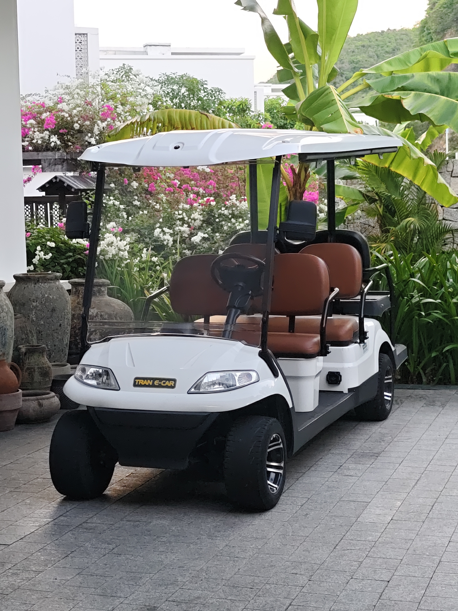 A white electric golf cart, labeled "TRAN E-CAR," is parked on a tiled pathway in what appears to be a resort or hotel setting. The cart has brown leather seats and a black steering wheel. In the background, there are lush green banana trees, vibrant pink and white bougainvillea flowers, and a large building with white walls and patterned architectural details. To the left of the cart, there are several large, weathered decorative pottery vases. The ground is paved with gray square tiles. The scene is outdoors, with natural light suggesting daytime. The location is identified as Bac Nha Trang Ward, Vietnam. There are no people visible in the image.