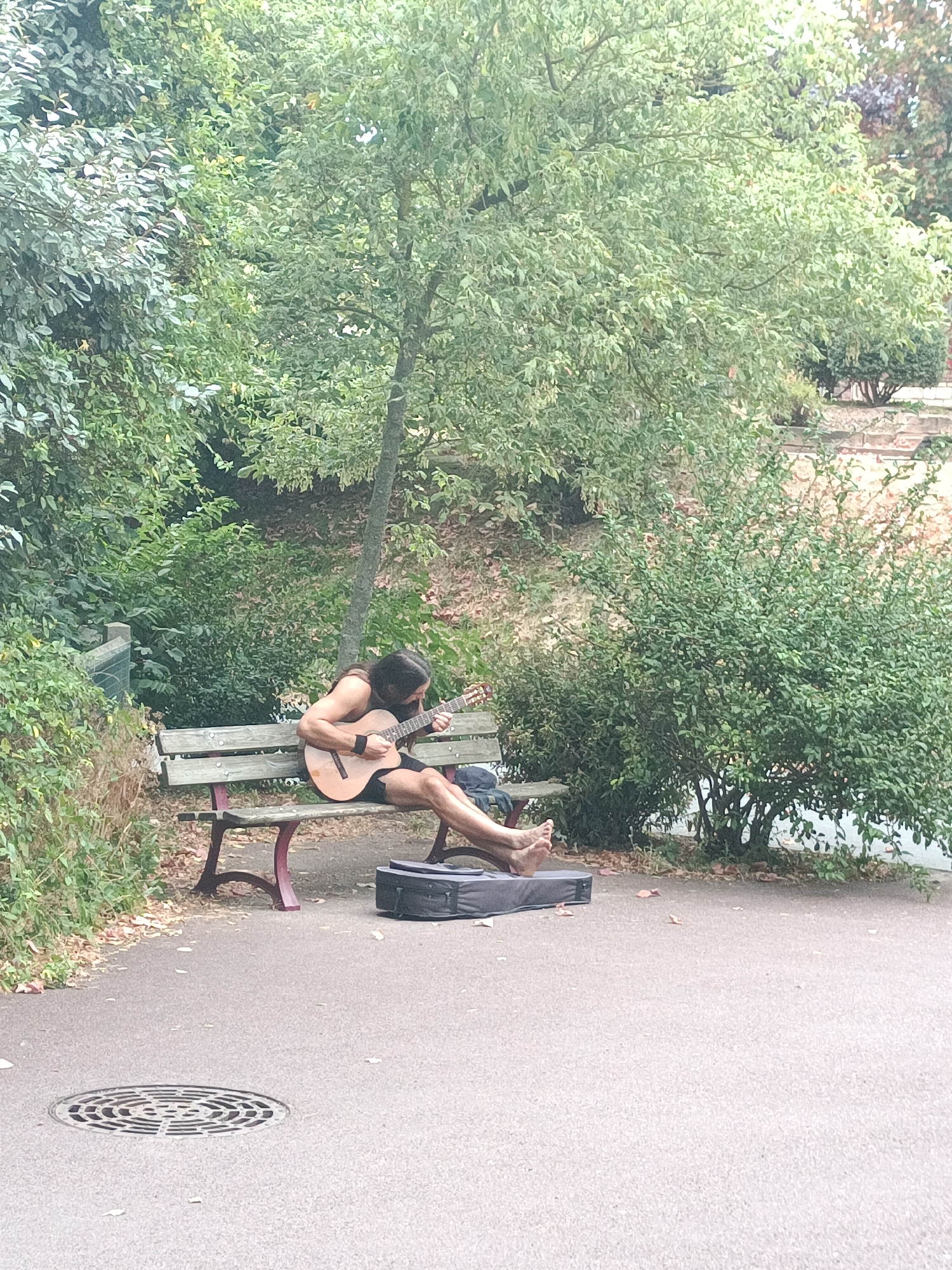 A long-haired, shirtless man with bare feet sits on a park bench and plays an acoustic guitar. A guitar case lies on the ground in front of him. The man is wearing dark shorts and a black wristband on his left wrist. He is sitting in a park with abundant green trees and bushes surrounding him. A paved walkway is in the foreground, with a circular grate visible on the ground. The overall impression is of a peaceful, outdoor scene in a park. This scene is located in the city of Houlgate, France.