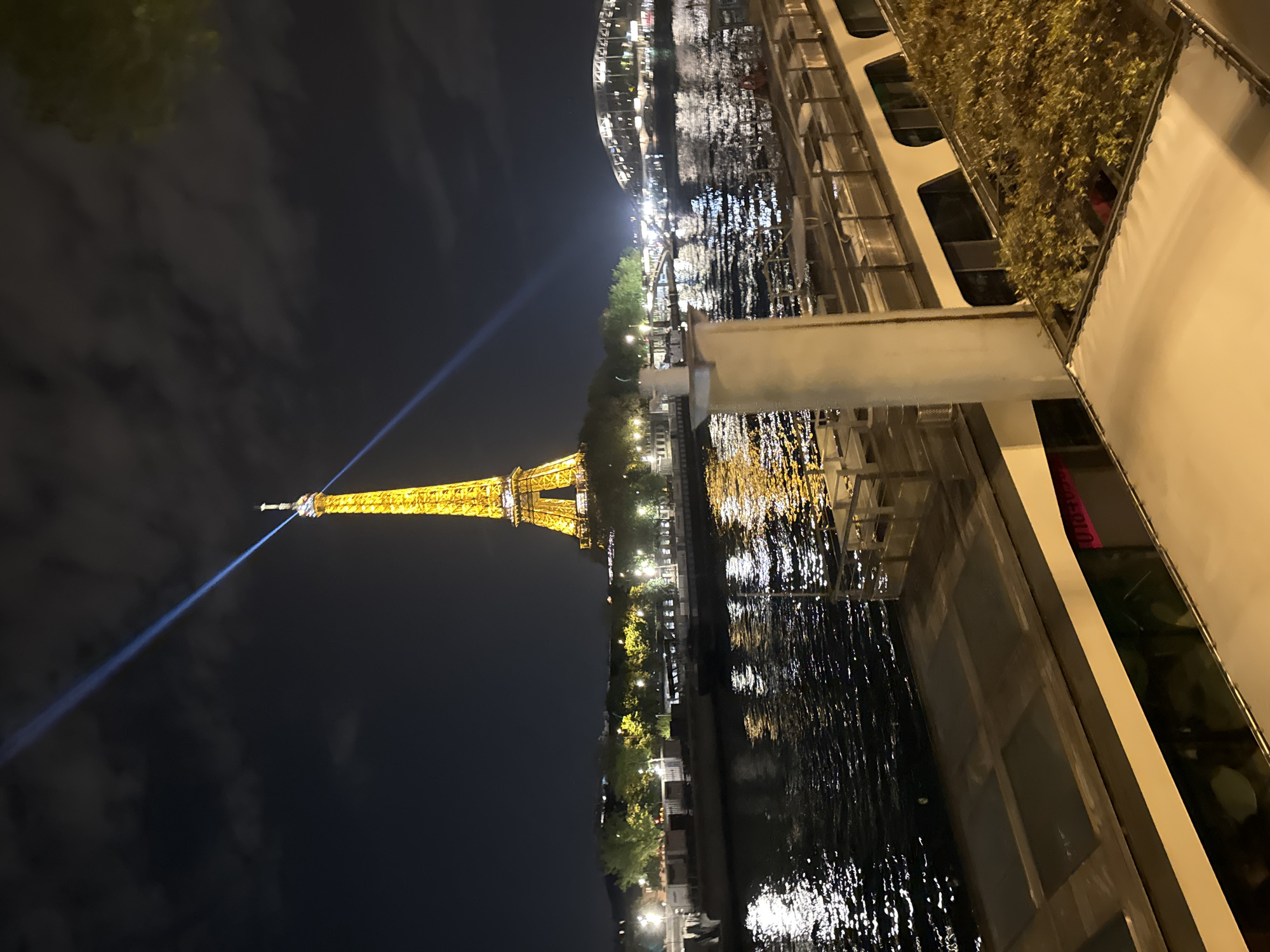 The image depicts the Eiffel Tower in Paris, France, at night. The tower is illuminated and its beam can be seen streaking into the dark sky. The view is from what appears to be a boat on a river, as the water reflects the city lights and parts of the boat's structure are visible in the foreground. There is no indication of people in the scene.