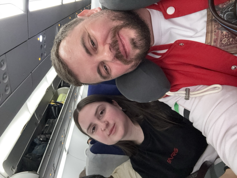 The image is a selfie taken inside an airplane cabin, featuring two individuals – a man and a woman – in a relaxed posture. The photo is oriented such that the subjects appear to be leaning to their left.
In the upper portion of the frame, a man with a beard and short dark hair is visible, smiling gently towards the camera. He is wearing a red jacket with white trim and white snap buttons, layered over a white t-shirt. A dark grey neck pillow is around his neck. Behind him, a patterned, possibly upholstered, surface can be seen.
Below and slightly to the left of the man is a woman with long dark hair, also looking towards the camera with a subtle, gentle expression. She is wearing a black t-shirt with the white letters "NRG" printed on the front. A dark grey neck pillow, similar to the man's, is around her neck.
The background clearly depicts the interior of an airplane. Above the subjects, a row of overhead storage bins is visible. Several of these bins are open, revealing various pieces of luggage and bags stored inside, including what appears to be a black backpack and a light green bag. The cabin interior is primarily dark grey, with bright artificial lighting strips running along the base of the overhead bins. Individual reading lights and air vents are also visible along the ceiling panel.
Both individuals appear comfortable and at ease, likely captured during a flight. The image conveys a personal moment of travel.
