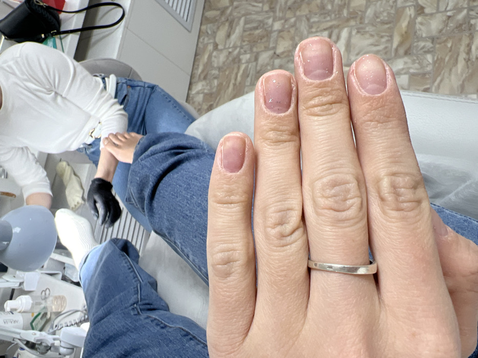 The scene depicts an interior view of a nail salon, reported to be located in Ust-Ilimsk, Russia. In the immediate foreground, a light-skinned left hand is presented, showcasing short, manicured nails with a translucent, light pink gel polish, some featuring small glitter particles. A plain silver band ring is worn on the fourth finger.
Behind the hand, the lower torso and legs of the client are visible, clad in blue denim jeans. The client's left foot, covered in a white ribbed sock, is elevated. Further in the background, a second individual, identified as a technician, is seated, wearing a white long-sleeved top, blue denim jeans, and black examination gloves. She is actively performing a pedicure on the client's bare right foot, which is positioned on a light-colored footrest. A gray, adjustable task lamp illuminates the work area.
To the left of the technician, on a white surface, various professional beauty products are arranged, including bottles (one partially labeled "ARAVIA"), an atomizer, and metal tools. The floor is covered in patterned light brown and beige tiles. A white wall or cabinet with horizontal vents is visible in the background, and a black handbag hangs on the right side of the frame. The environment appears brightly lit and sterile.