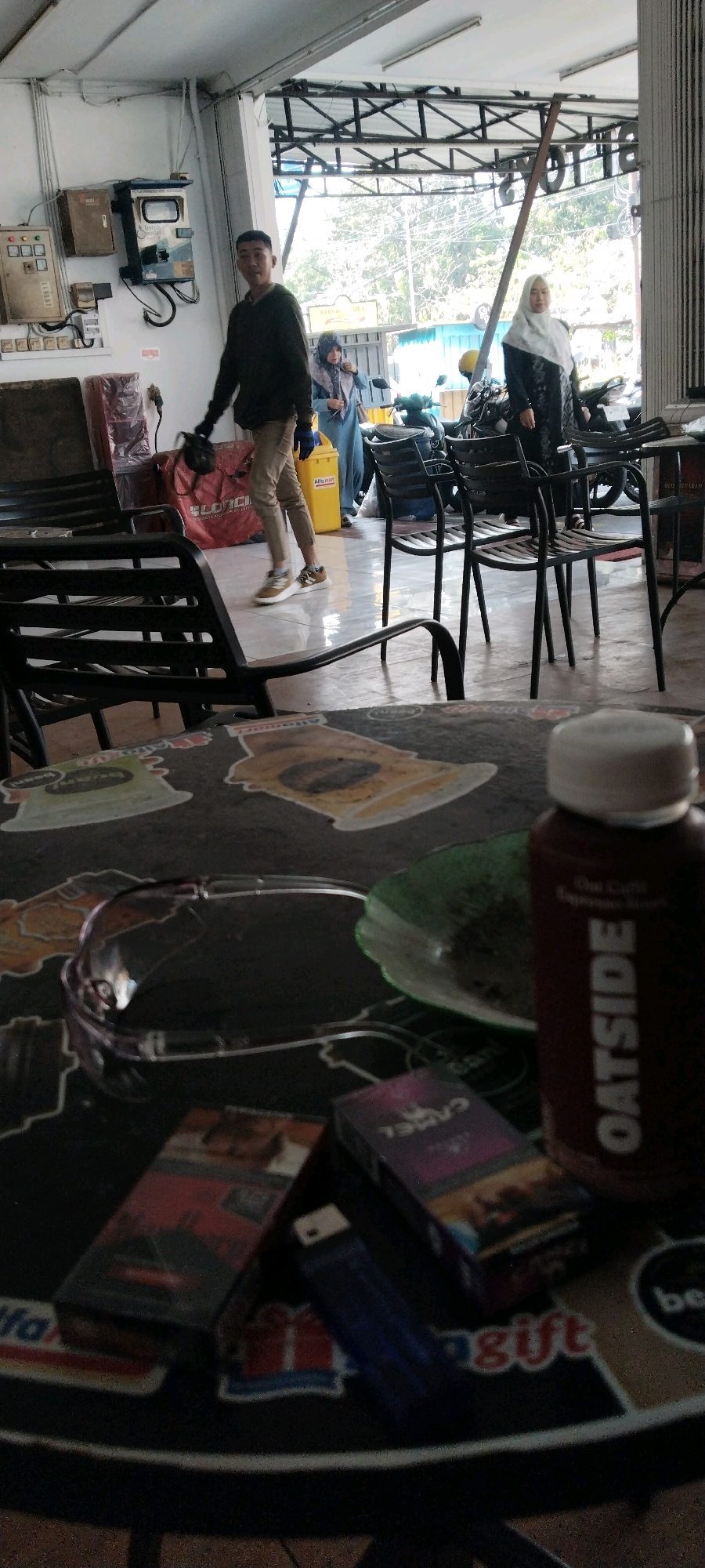 The image depicts an indoor scene, likely a cafe or open-air establishment, with a foreground focus on a table laden with various items. A clear plastic pair of safety glasses rests on the patterned table surface. Adjacent to the glasses is a green, translucent plastic plate. To the right of these items is a dark brown bottle with white lettering that reads "OATSIDE." In front of the bottle, two packs of cigarettes are visible: one red and gold, and another purple and gold with the brand name "CAMEL" printed on it. A blue lighter is positioned between the cigarette packs.
The background of the image reveals a man in a dark long-sleeved shirt and beige pants, walking towards the left. He is carrying a black helmet and wearing gloves. Further into the background, two women are present. One woman, wearing a light blue hijab and matching outfit, is looking towards the right. Another woman, wearing a dark hijab and dark patterned dress, is standing near a motorcycle. Several chairs and tables are arranged in the area, with some motorcycles parked in the vicinity. The environment appears to be an open-air seating area with a roof structure overhead. Electrical conduits and boxes are visible on a white wall in the background. The overall impression is a casual, public gathering space.