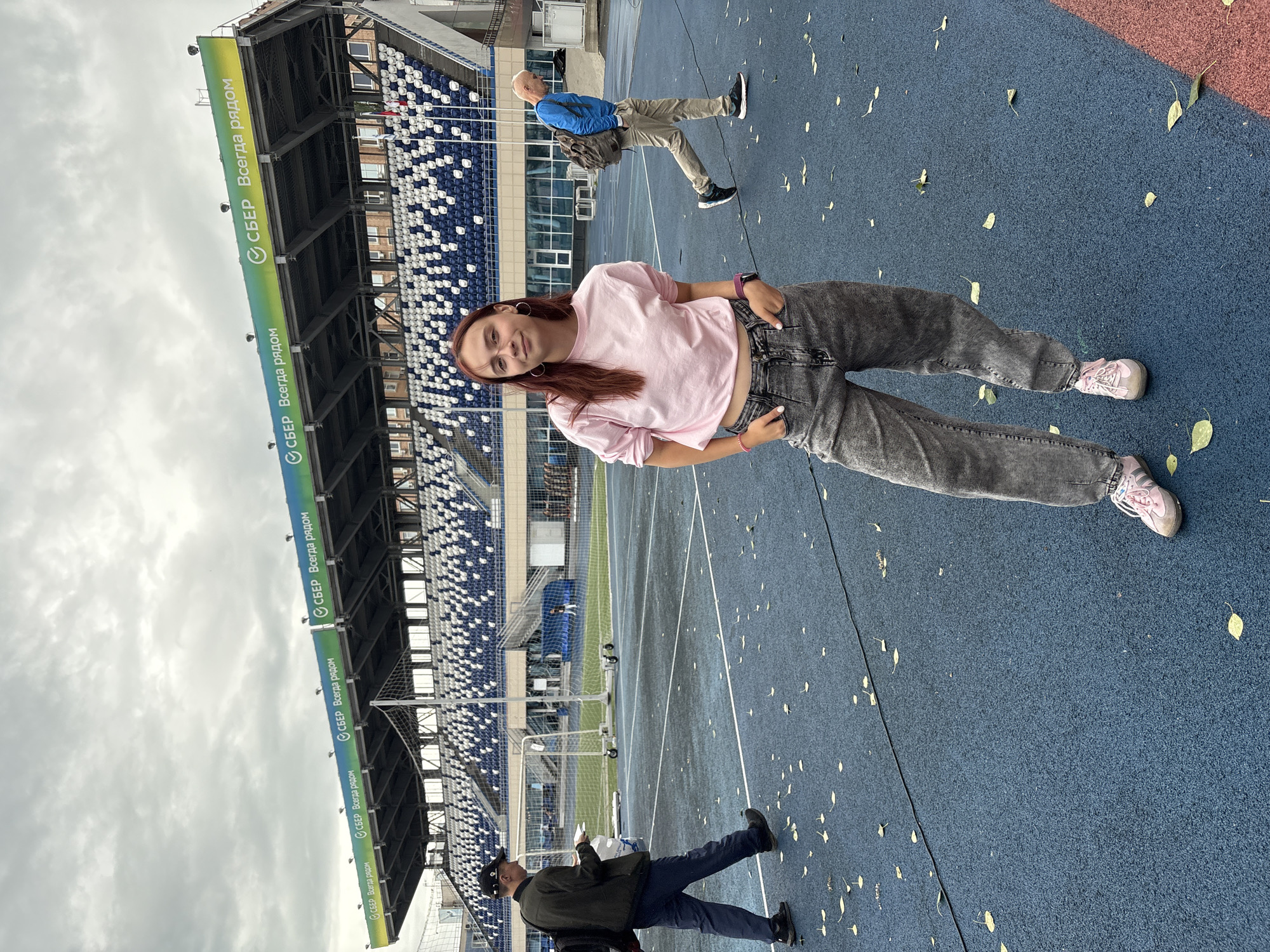 This media file shows a full-body shot of a young woman standing in an outdoor stadium. She is wearing a light pink t-shirt and grey, baggy jeans, with pink sneakers. She is looking at the camera with a slight smile. Behind her, an older man in a blue jacket and beige pants is walking away. The stadium features blue and white seating in the background, with a blue track and field area in the foreground. The weather appears overcast, and there are scattered yellow leaves on the ground. Along the stadium's upper structure, there is visible text that reads "СБЕР Всегда рядом" in Russian, which translates to "Sber Always Nearby," indicating sponsorship. The scene is set in a stadium, likely for sports or athletic events, in Izhevsk, Russia.
