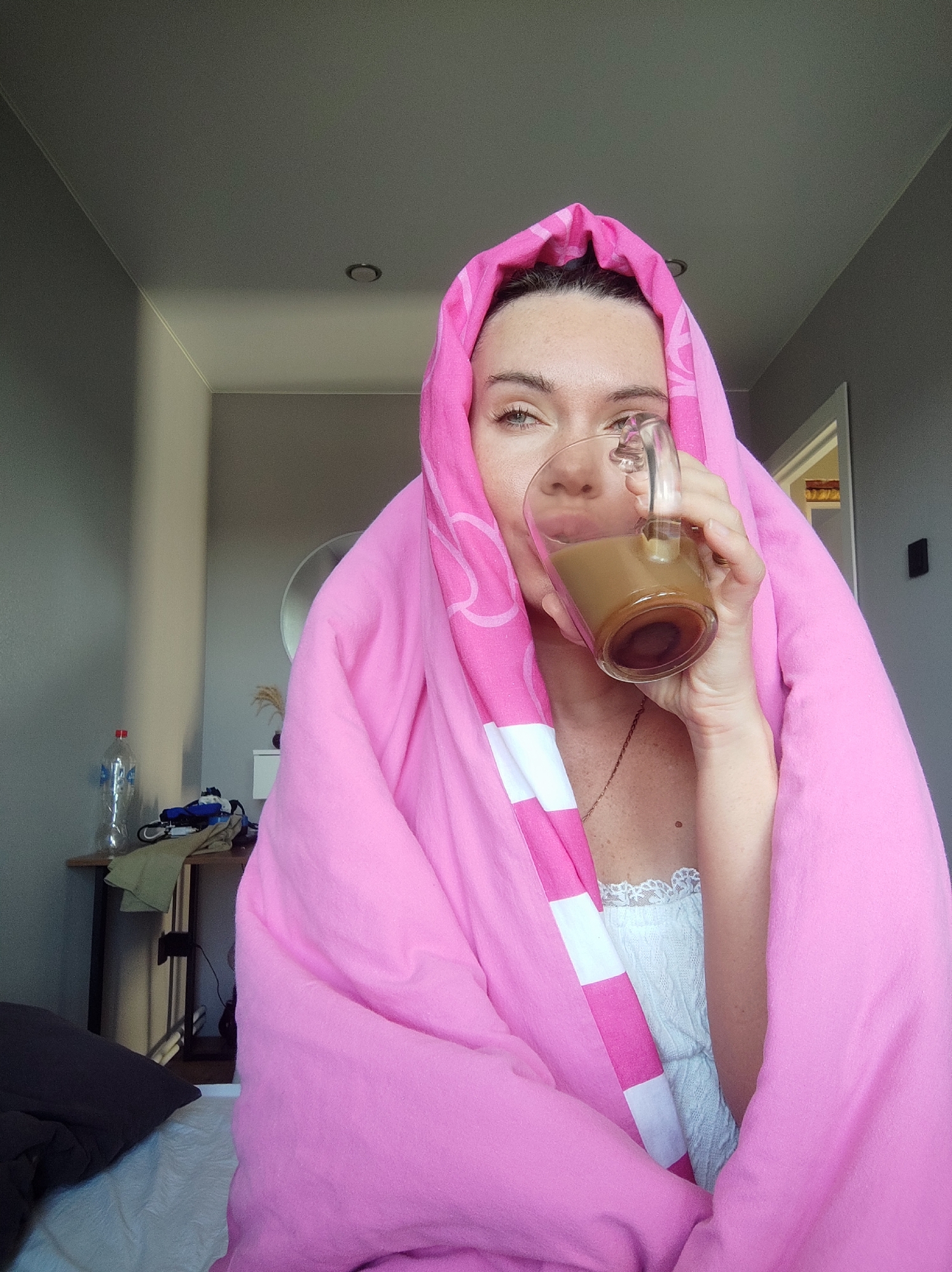 A person wrapped in a pink and white striped towel is holding a glass mug of coffee in their right hand. The person has light skin, brown hair, and light eyes. The mug is transparent, revealing a brown liquid. The person is wearing a white lacy top underneath the towel. The location appears to be indoors in a residential setting. There is a bed with a pillow in the lower left of the frame, a table with miscellaneous items in the background to the left, and a doorway to another room to the right. The walls are painted a neutral grey color. There are no indications in the image that the location is specifically in Votkinsk, Russia.