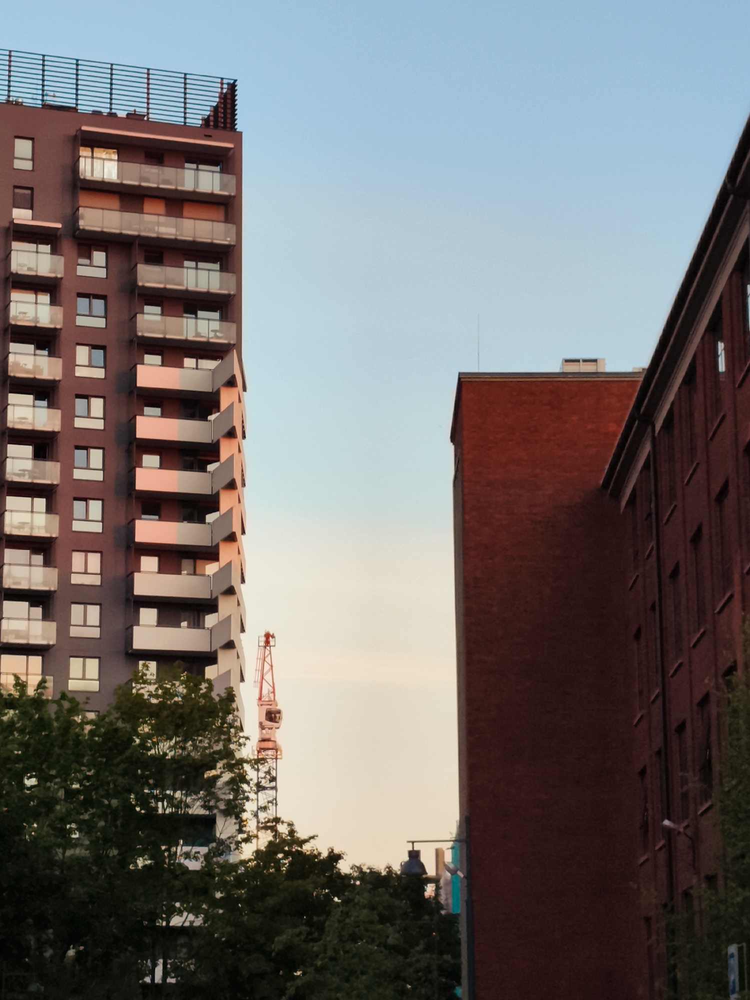 This is a photograph of an urban cityscape, likely in Gdańsk, Poland, captured during twilight. The image is dominated by two modern apartment buildings and a construction crane.
The building on the left is a tall, slender residential tower with a dark facade and numerous balconies. The balconies have glass railings and are accented with light-colored panels. The building on the right is a lower, wider structure made of red brick, with a series of windows.
A red construction crane stands prominently in the middle ground, its boom extending upwards. In the foreground, a dense canopy of green trees partially obscures the view of the lower parts of the buildings. The sky is a pale blue, transitioning to a warmer hue near the horizon, indicating either sunrise or sunset. There are no visible people or animals in the frame. The overall impression is one of urban development and construction.