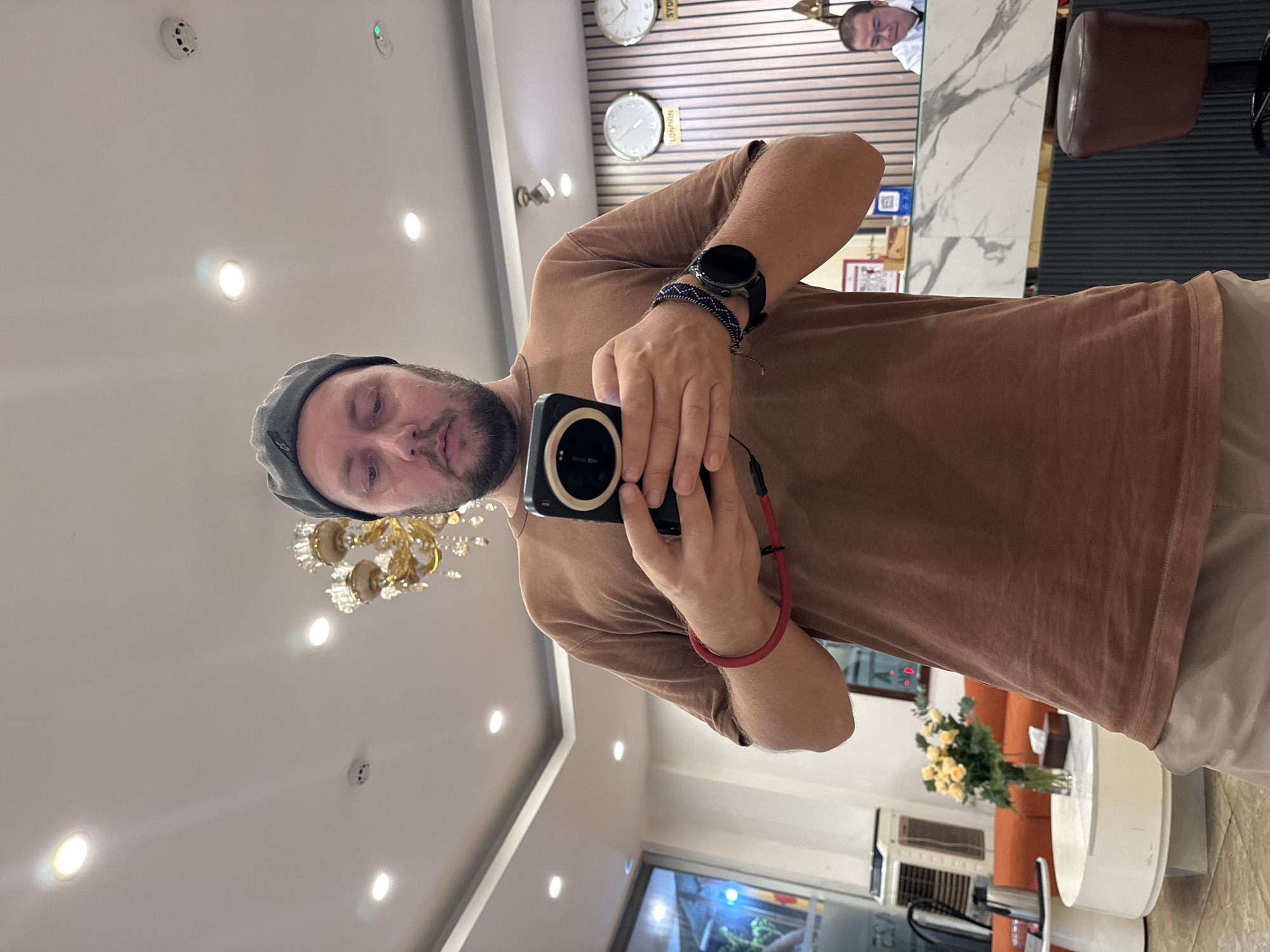 This is a vertical, first-person photo taken by a man holding a camera to take a selfie. The man is wearing a grey cap, a brown t-shirt, and tan pants. He has a beard and appears to be looking at the camera. On his left wrist, he wears a smartwatch. The background of the image shows a hotel lobby. On the wall behind him, there are multiple clocks showing different times, including one labeled "LONDON" and another labeled "SYDNEY." A chandelier hangs from the ceiling. In the reflection of a marble countertop, another man wearing glasses and a white shirt can be seen. To the right of the frame, a brown leather chair is visible. Further in the background, a bouquet of yellow flowers in a glass vase sits on a table. The scene appears to be indoors, and given the lighting, it's likely daytime or late evening. There is a visible QR code on the wall behind the man, and some text on a sign below it. The man's expression is neutral, and the overall tone of the image is casual.
