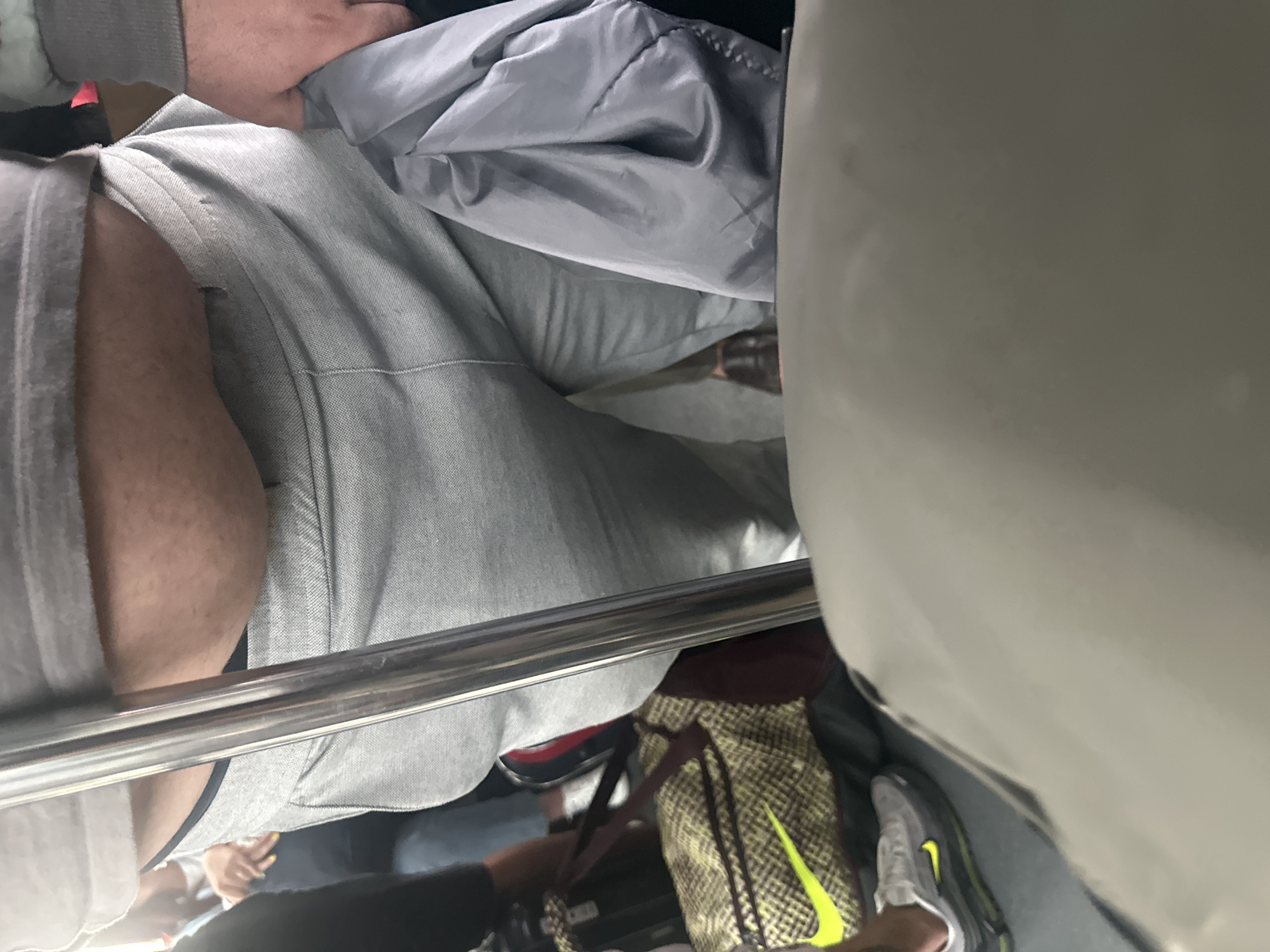 An individual is standing on a public transport vehicle, possibly a bus or train, evidenced by the metal pole in the foreground and other passengers in the background. The individual is wearing grey sweatpants and a grey t-shirt, with a portion of their midsection visible between the two garments. A dark-colored garment is draped over their left arm, and a patterned duffel bag with a neon yellow Nike logo is visible on the floor to their left. Their feet are clad in dark-colored shoes. The lighting suggests an indoor environment.