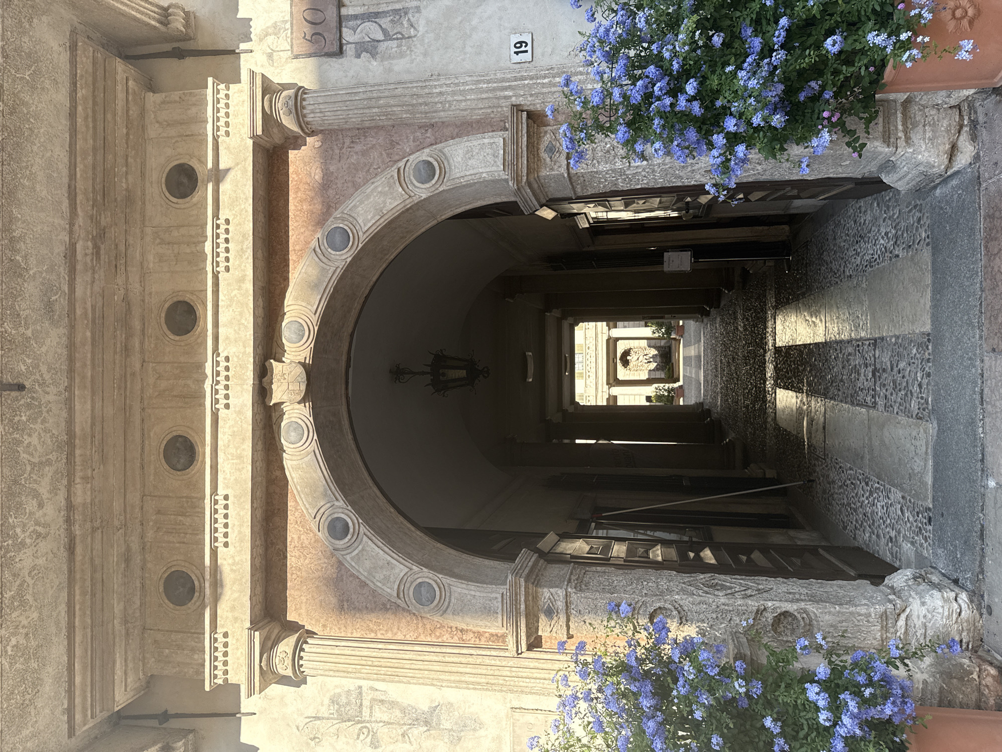 The image depicts a narrow, arched passageway bathed in sunlight, suggesting a clear day. The architecture is classical, with ornate columns, stone carvings, and circular decorative elements visible on the facade. The passageway leads to a courtyard or another open area, where a statue or fountain is partially visible in the distance. The ground within the passageway is paved with stones, with alternating patterns of light and dark, indicating a textured surface.
Blue flowers cascade from planters on the right side of the image, adding a touch of vibrant color to the scene. The overall impression is one of historical architecture and serene beauty. There is a visible number "19" on a white plaque on the upper wall, and "50" is also visible on another plaque. The lighting suggests it is either morning or afternoon, with strong shadows cast by the architectural elements. The scene appears quiet and peaceful, with no people or animals visible, focusing solely on the architectural details and the natural element of the flowers. The stone texture and the presence of classical architectural styles suggest an older, possibly European, urban setting. Given the location context of Trento, Italy, this is likely a historical part of the city.